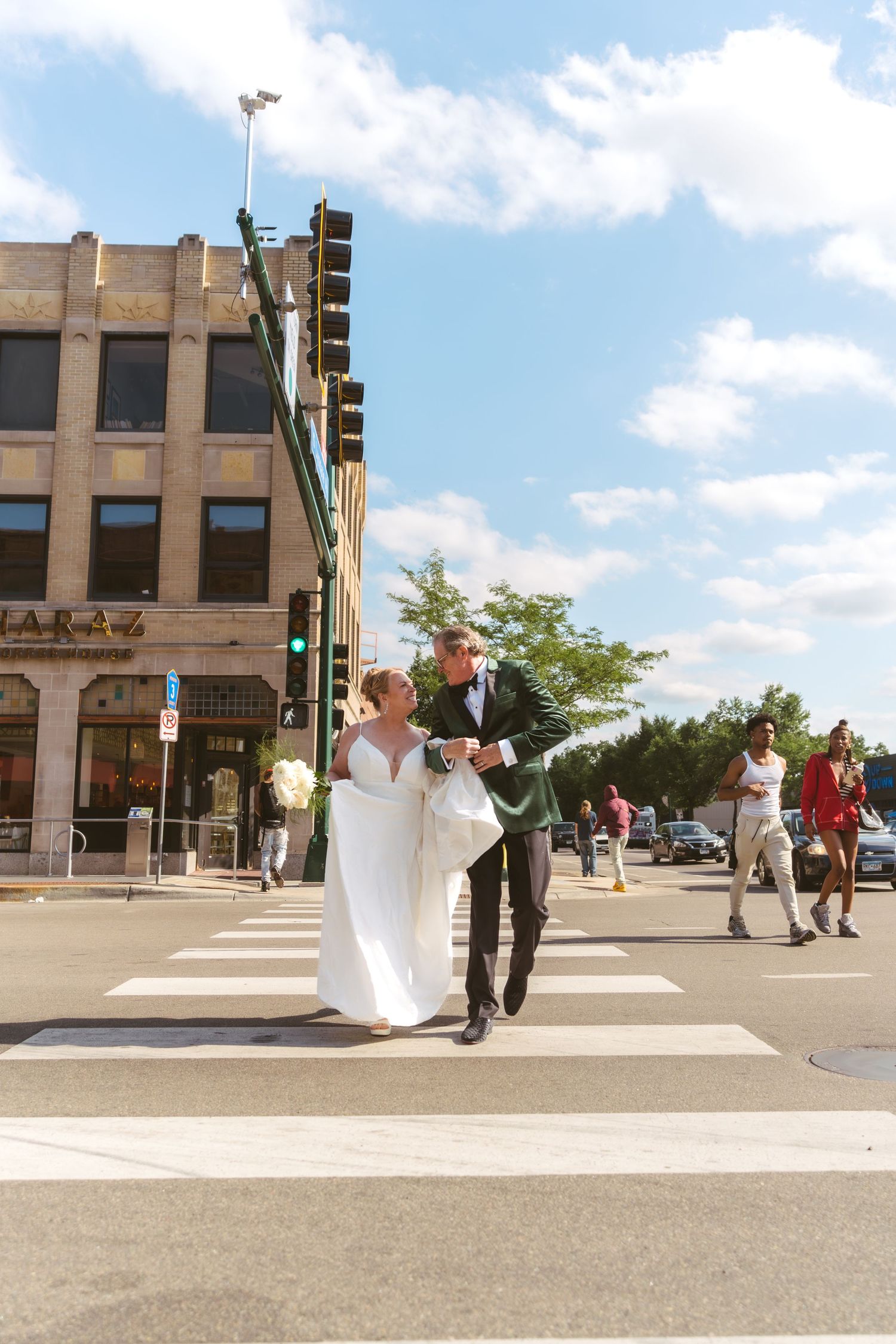 Bride and groom cross a busy urban intersection together on their wedding day under blue skies.