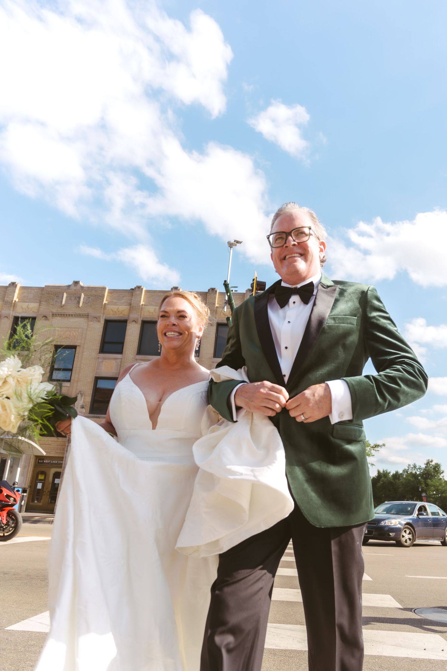Smiling bride and groom in green tuxedo pose outdoors under blue sky in a city street setting.