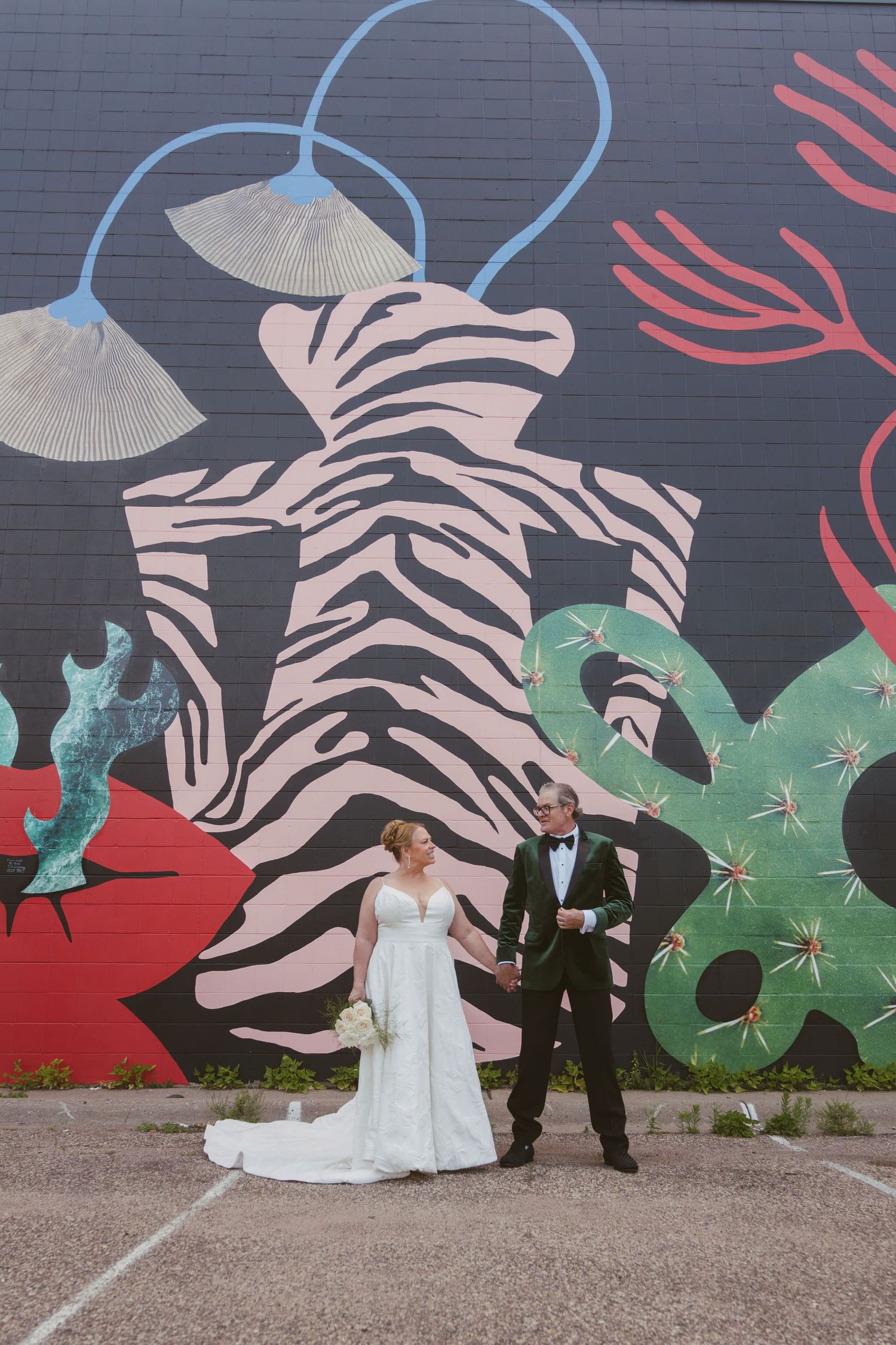 Bride and groom hold hands before a vibrant colorful zebra mural, bride in white gown with bouquet.