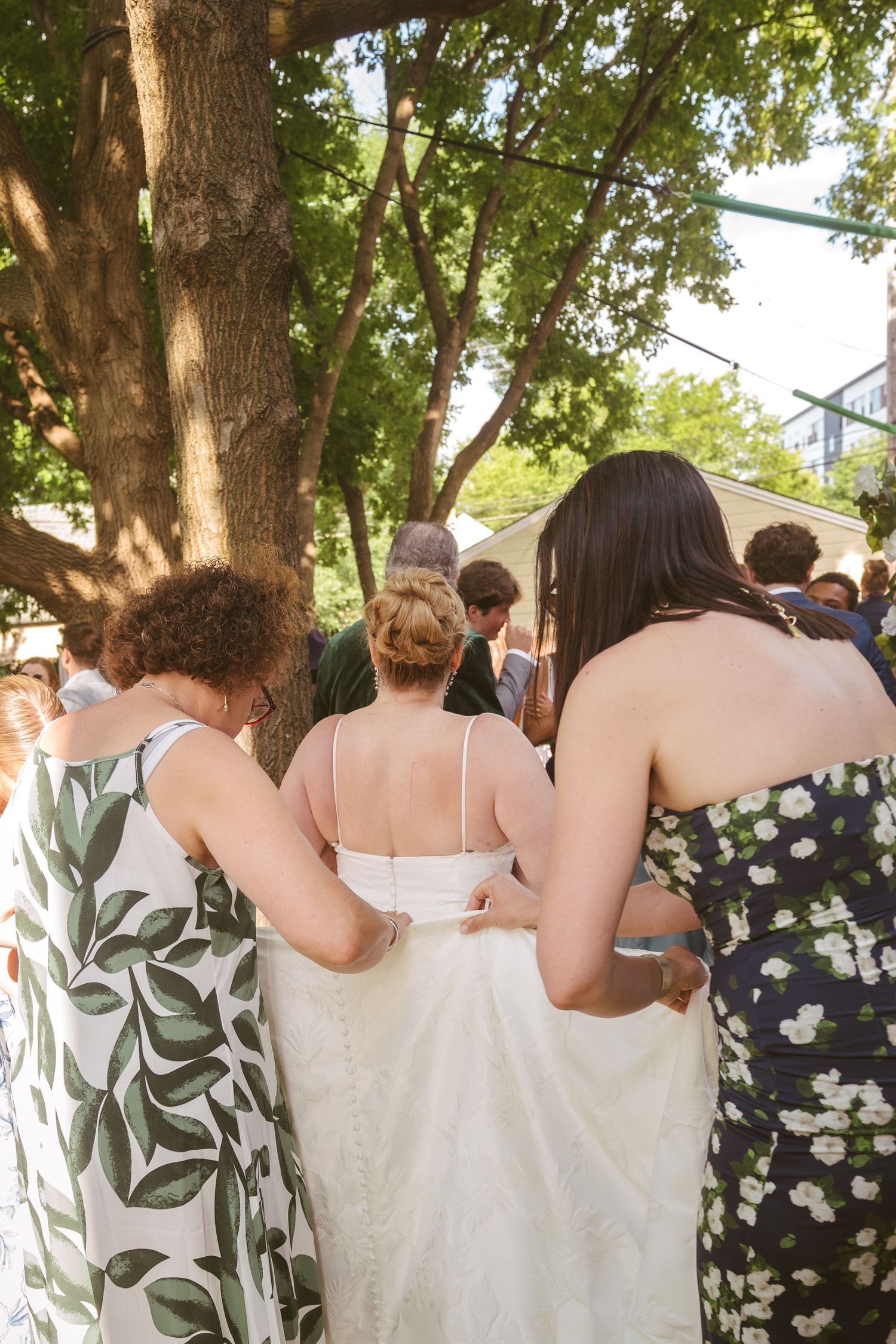 Bridesmaids helping bride with wedding dress outdoors under trees.