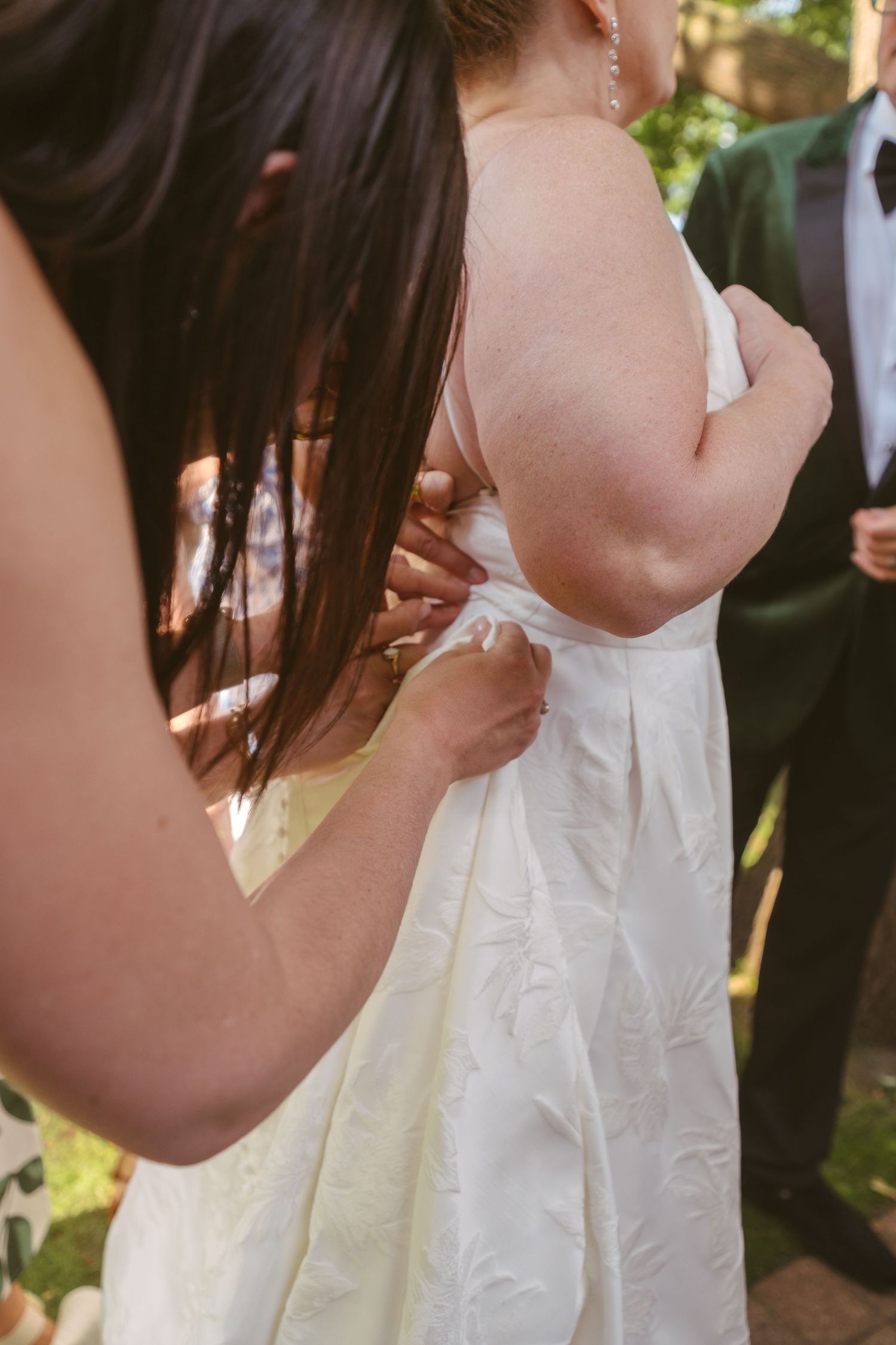 A bridesmaid with dark hair fastens the back of the bride's white floral embossed wedding gown outdoors.