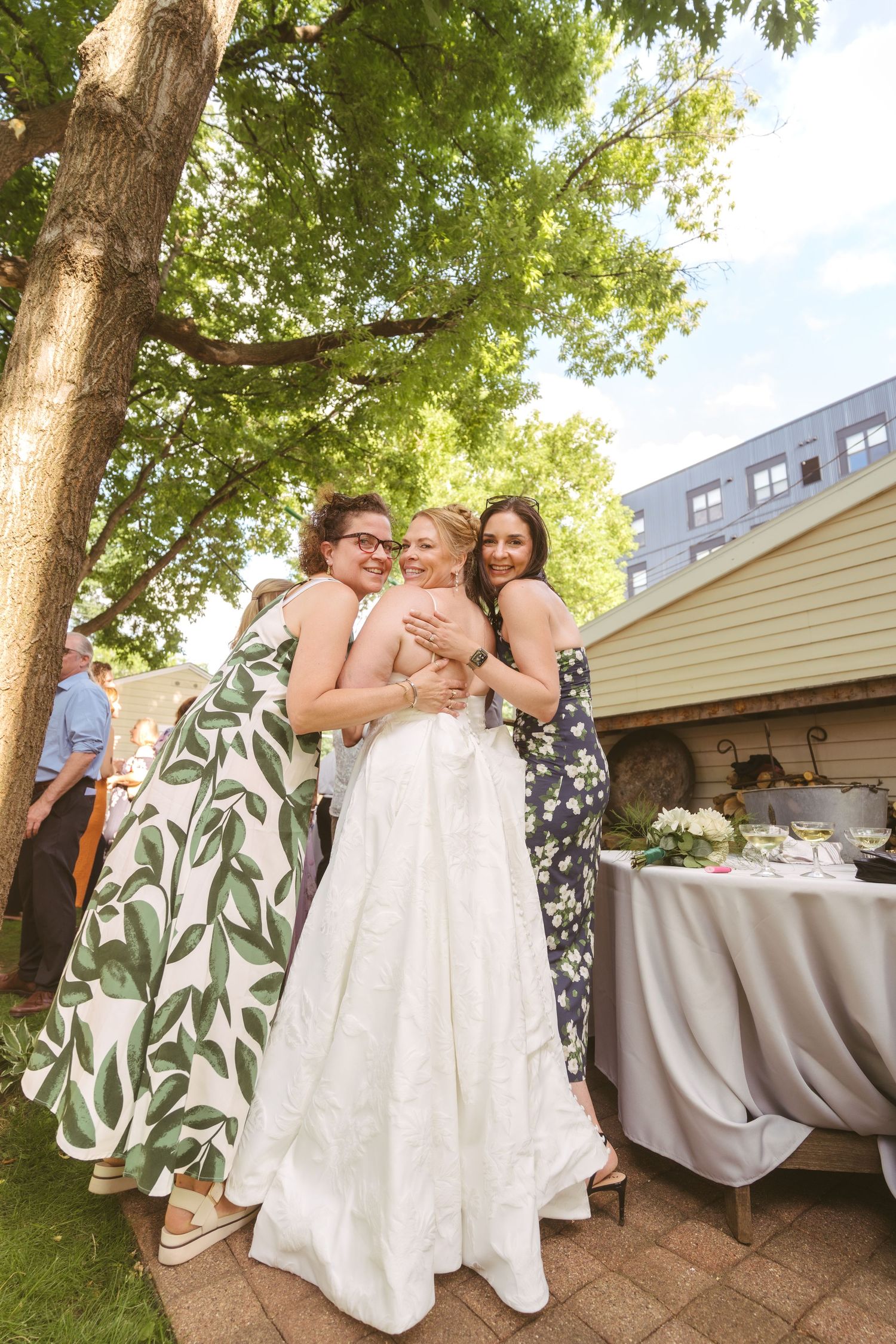 Bride in white gown receives warm hugs from two guests during outdoor wedding reception.