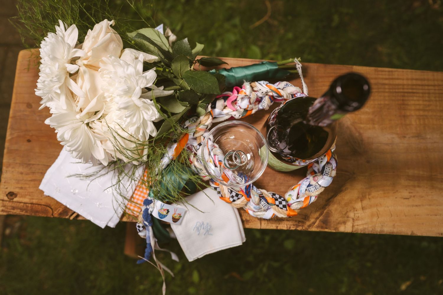 Rustic wooden bench with white peony bouquet, wine glass, and bottle decorated with colorful ribbon outdoors.