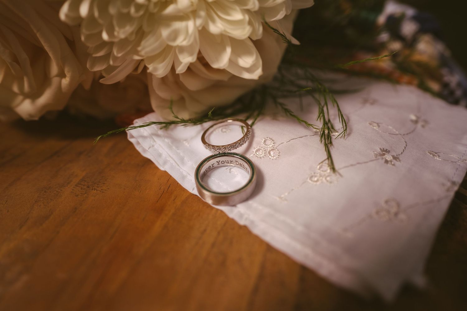 Two wedding bands rest on embroidered white linen beside a white peony bloom on a wooden surface.