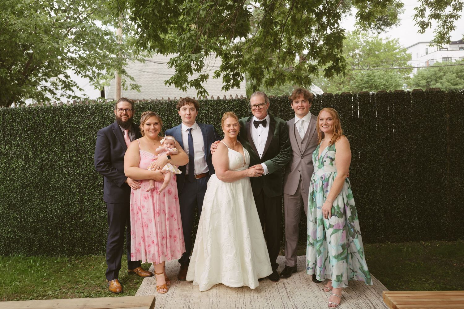 Wedding party of seven posing outdoors in front of lush green hedge, bride in white gown centered among formally dressed guests.