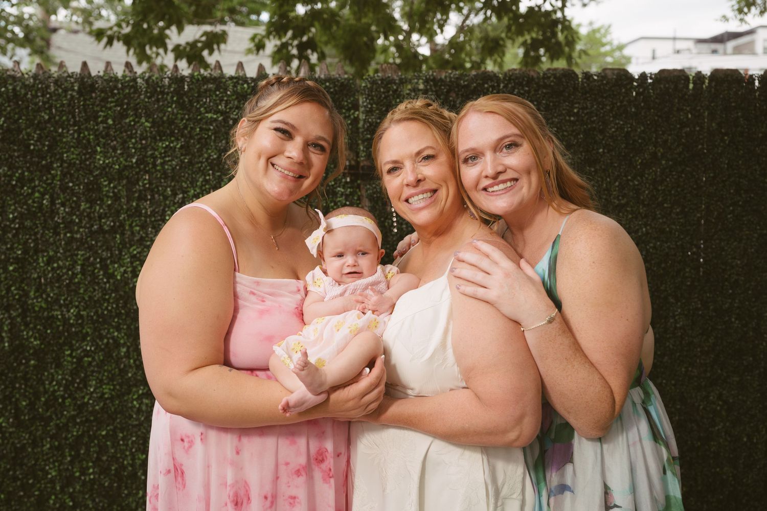 Three smiling women pose outdoors holding a baby girl in a pink headband near a green hedge.
