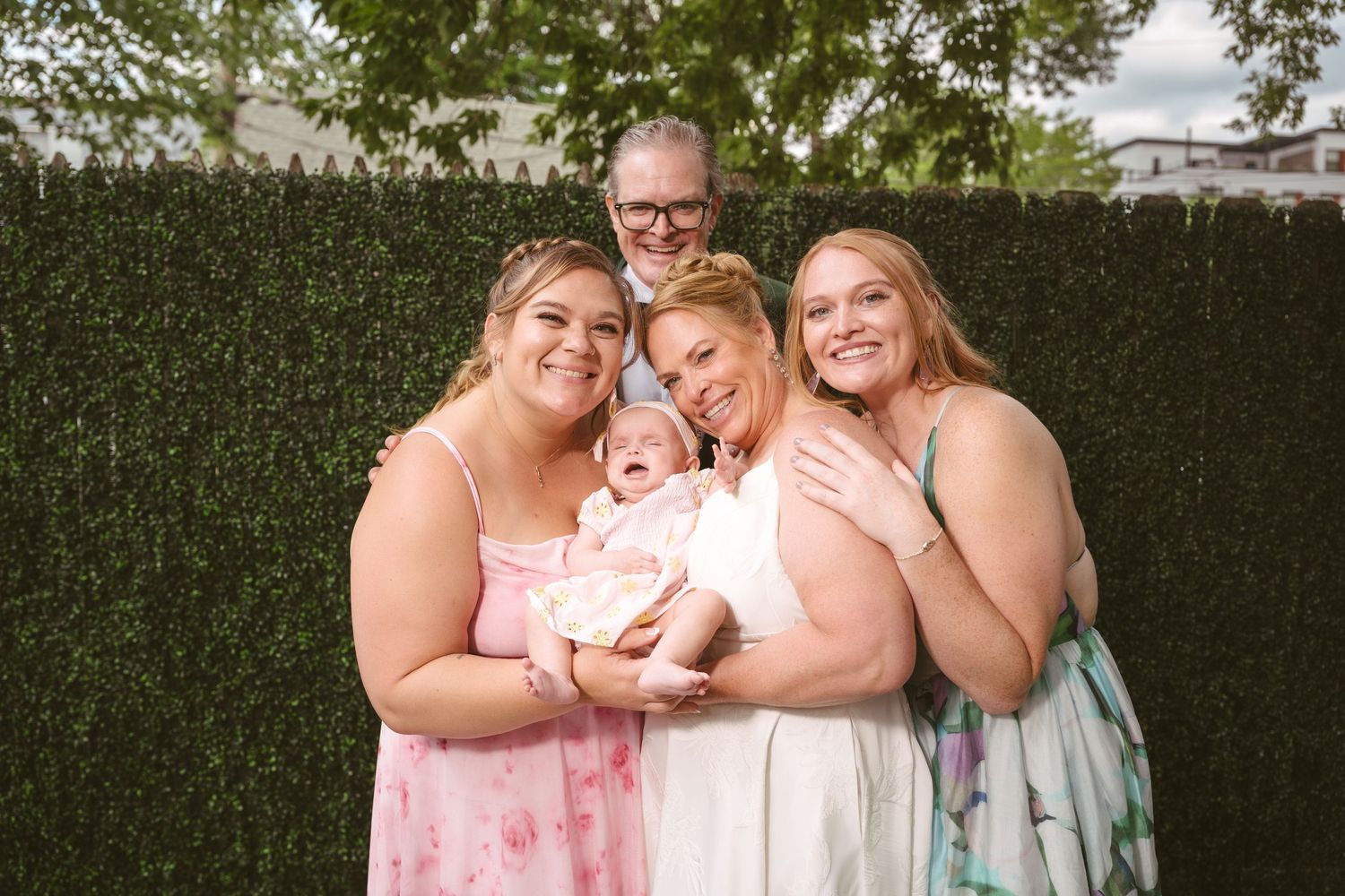 Family portrait with parents and two adult daughters holding a baby in pink outdoors near a green hedge wall.