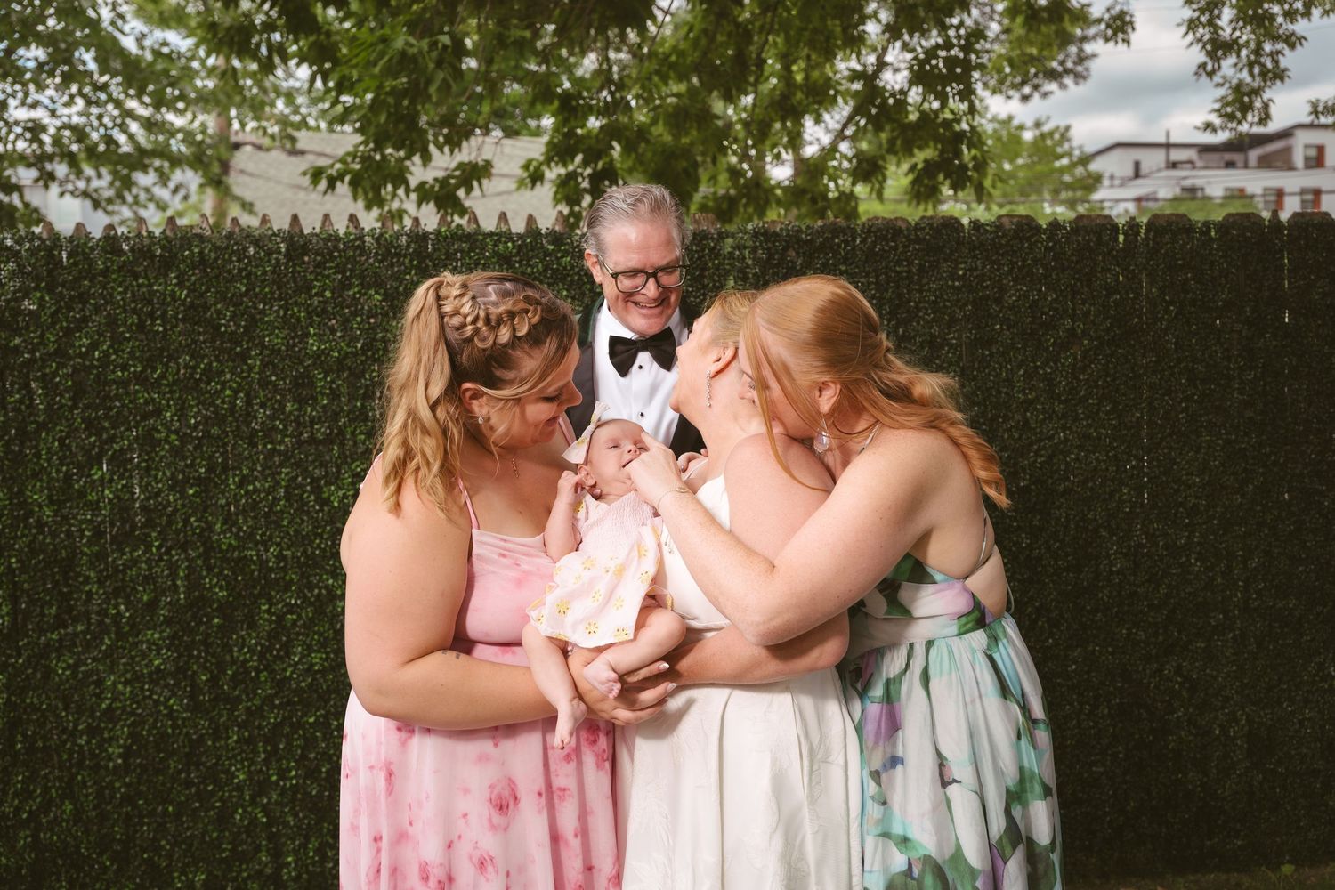 Family gathering outdoors near green hedge, two women in floral dresses admiring baby while man in tuxedo smiles behind them.