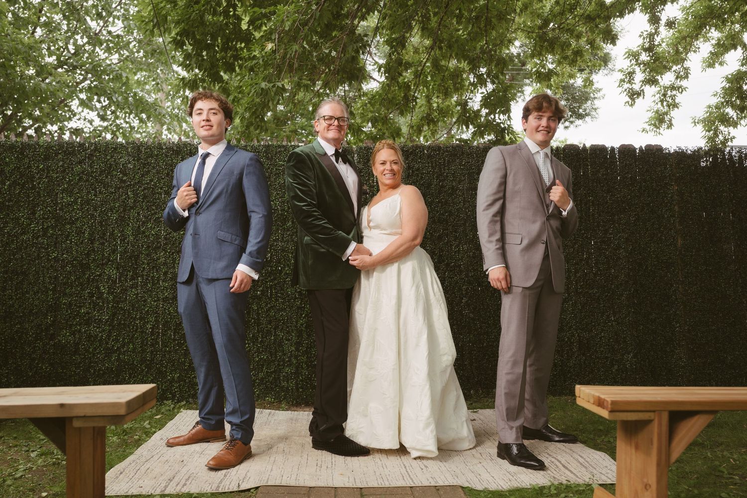 Wedding party poses outdoors with hedgerow backdrop, bride in white gown flanked by groom and groomsmen in suits.