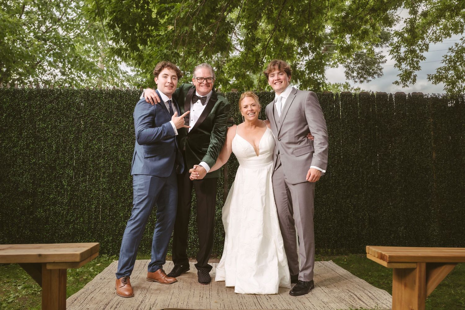Bride in white gown poses with three men in suits and tuxedo outdoors at wedding reception near green hedge.