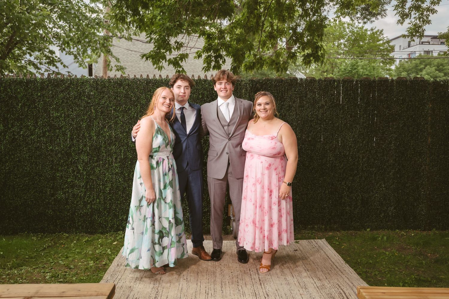 Four guests in formal attire pose together outdoors at a wedding reception with lush green hedge background.