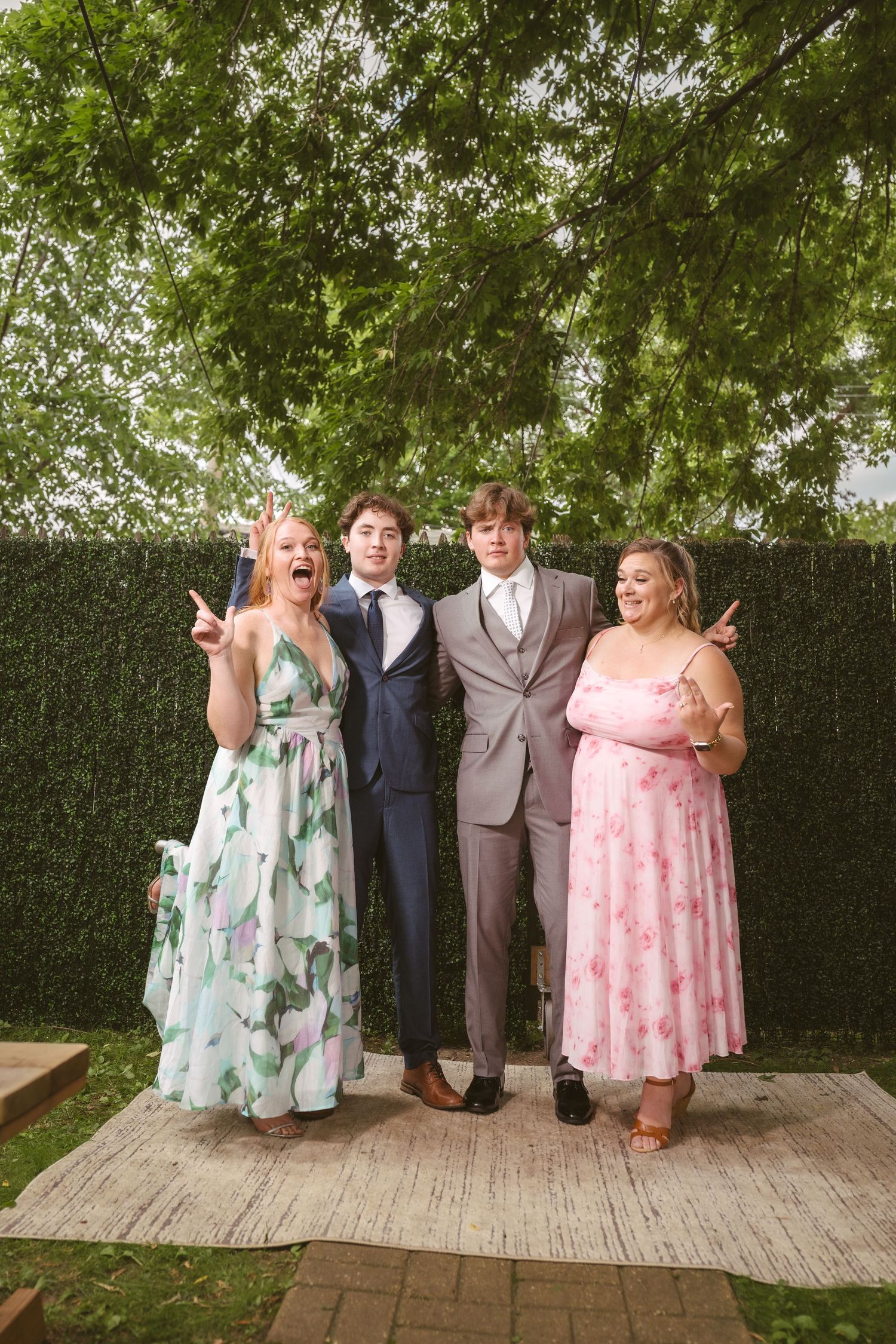 Two young couples pose playfully outdoors in formal attire, wearing prom dresses and suits against a lush green hedge backdrop.