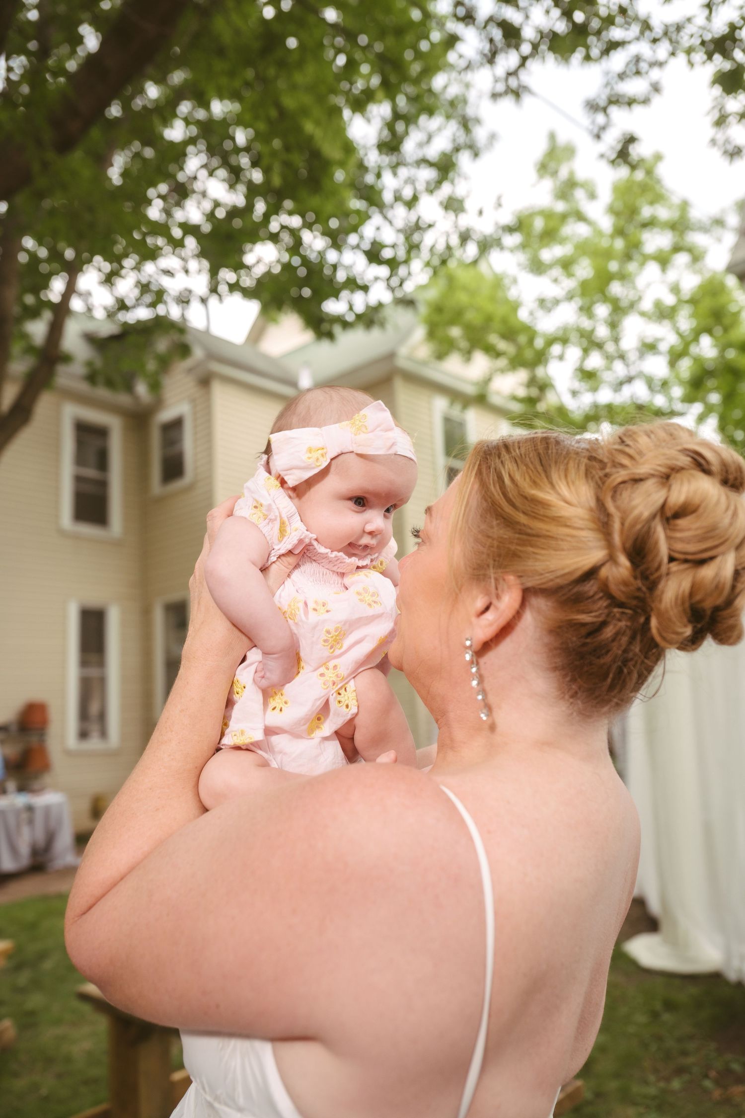 Mother in white dress holds baby in pink floral outfit up to her face outdoors near a house.