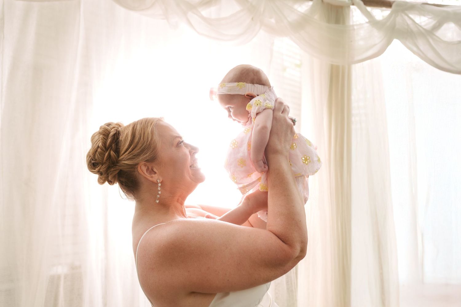 Bride in white gown lifts smiling baby girl in soft natural light near sheer curtained window.