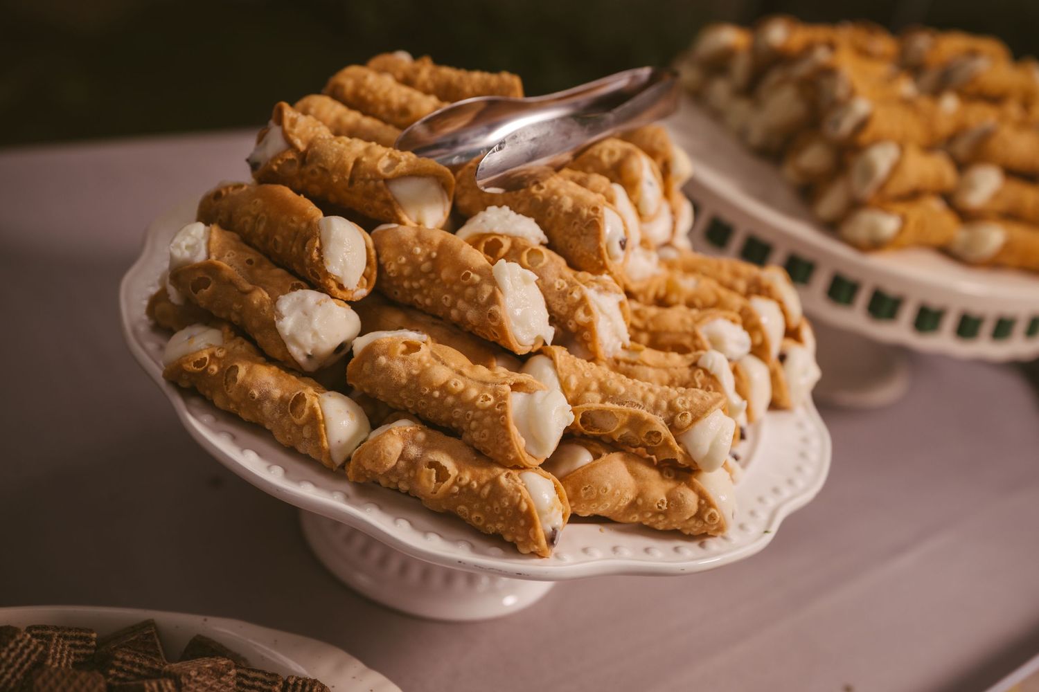 White pedestal dish piled high with golden cannoli pastries filled with cream, displayed at a wedding reception dessert table.