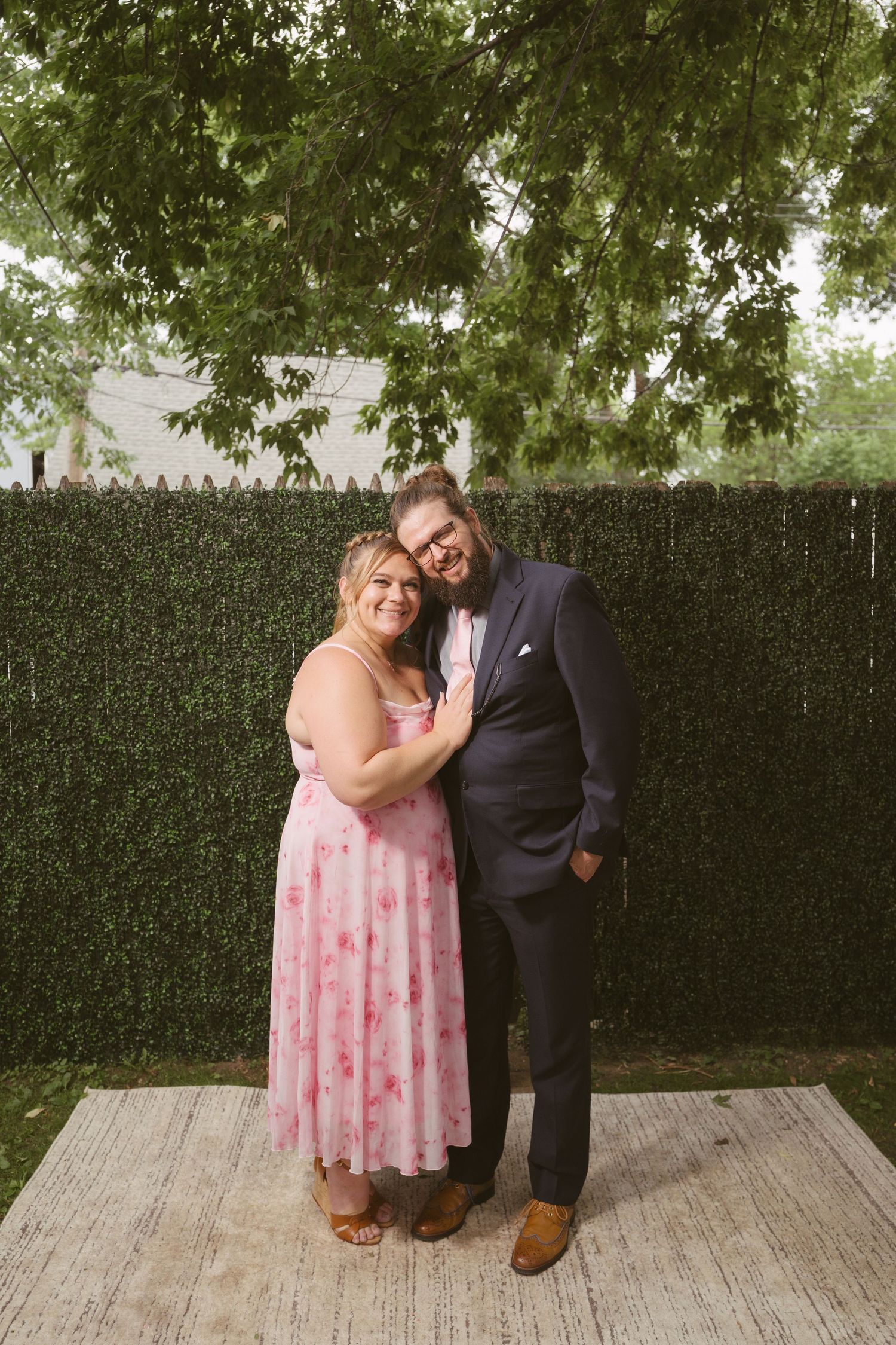 Couple posing together outdoors, woman in pink floral dress and man in navy suit, smiling in front of green hedge backdrop.