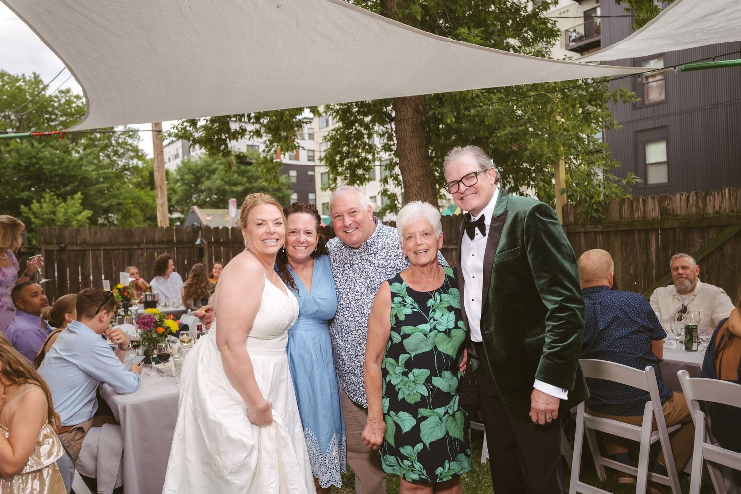 Group of five people posing together at an outdoor wedding reception, with bride in white gown.