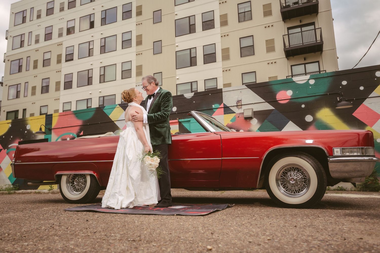 Bride and groom kissing beside a vintage red Cadillac convertible in front of a colorful urban mural.