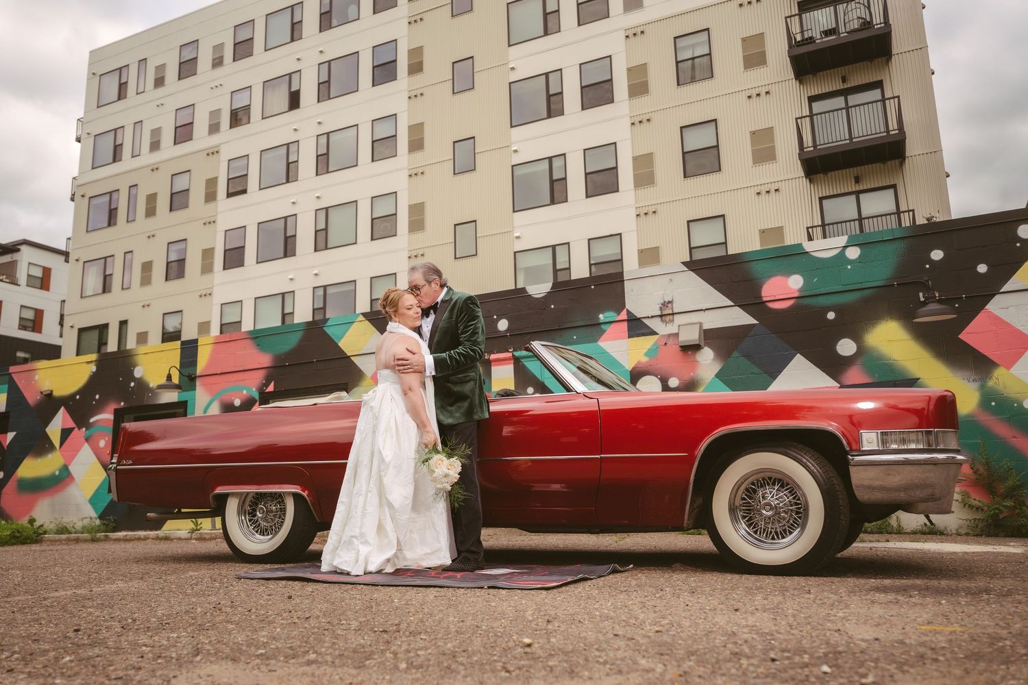 Newlyweds kissing beside a vintage red Cadillac with a colorful mural backdrop.