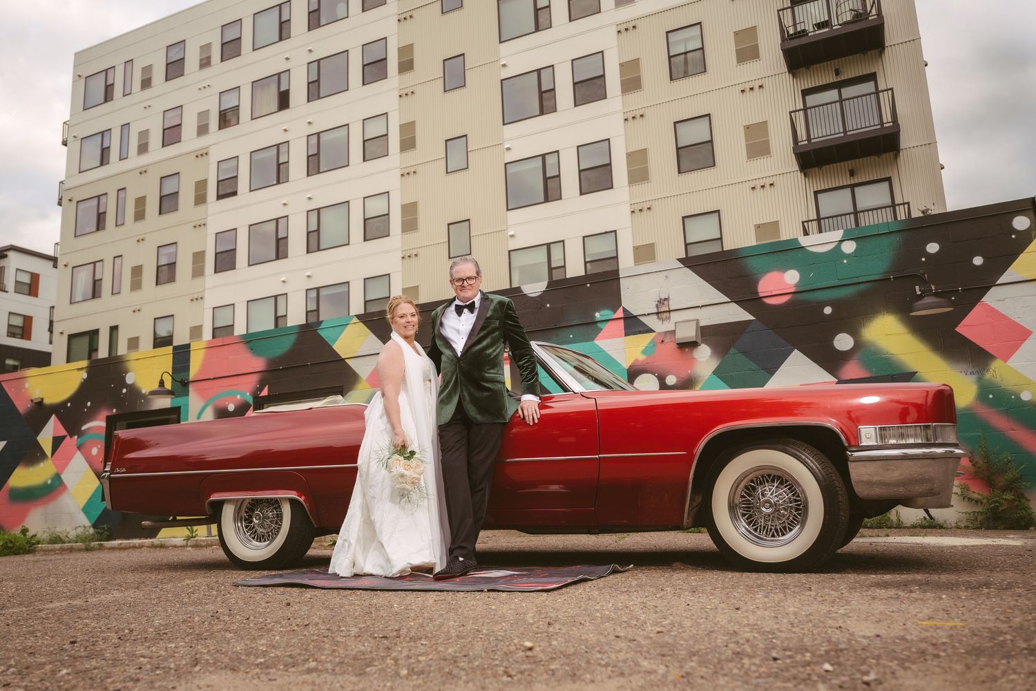 Bride and groom pose beside a classic red vintage Cadillac convertible in front of a colorful mural.
