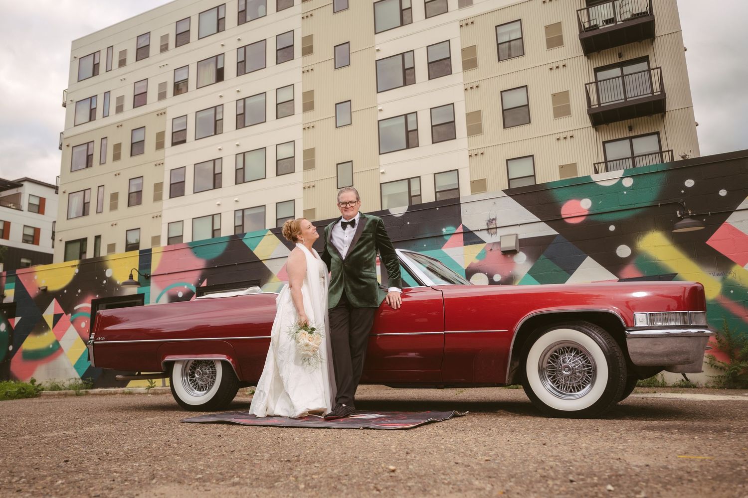 Bride and groom posing by a classic red convertible Cadillac in front of a vibrant street mural.