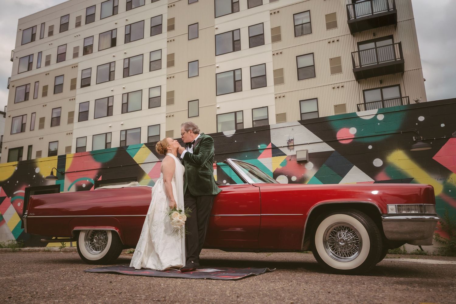 Bride and groom share a kiss beside a classic red Cadillac with a colorful urban mural backdrop.