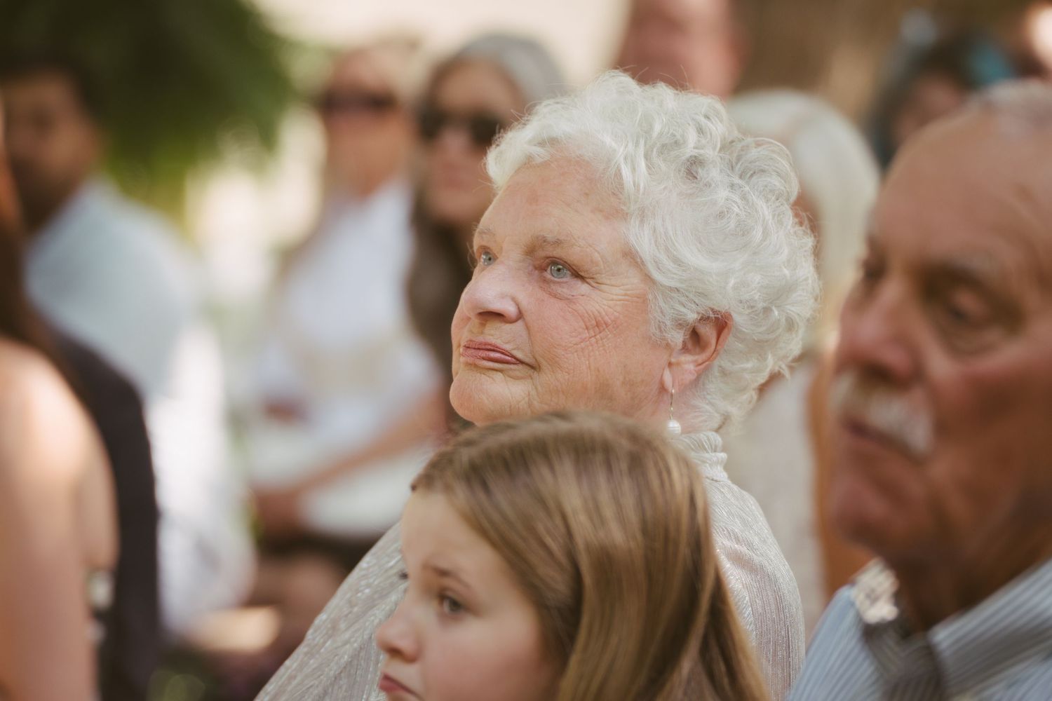 Elderly white-haired woman watches emotionally as a young girl sits in front of her at an outdoor wedding ceremony.