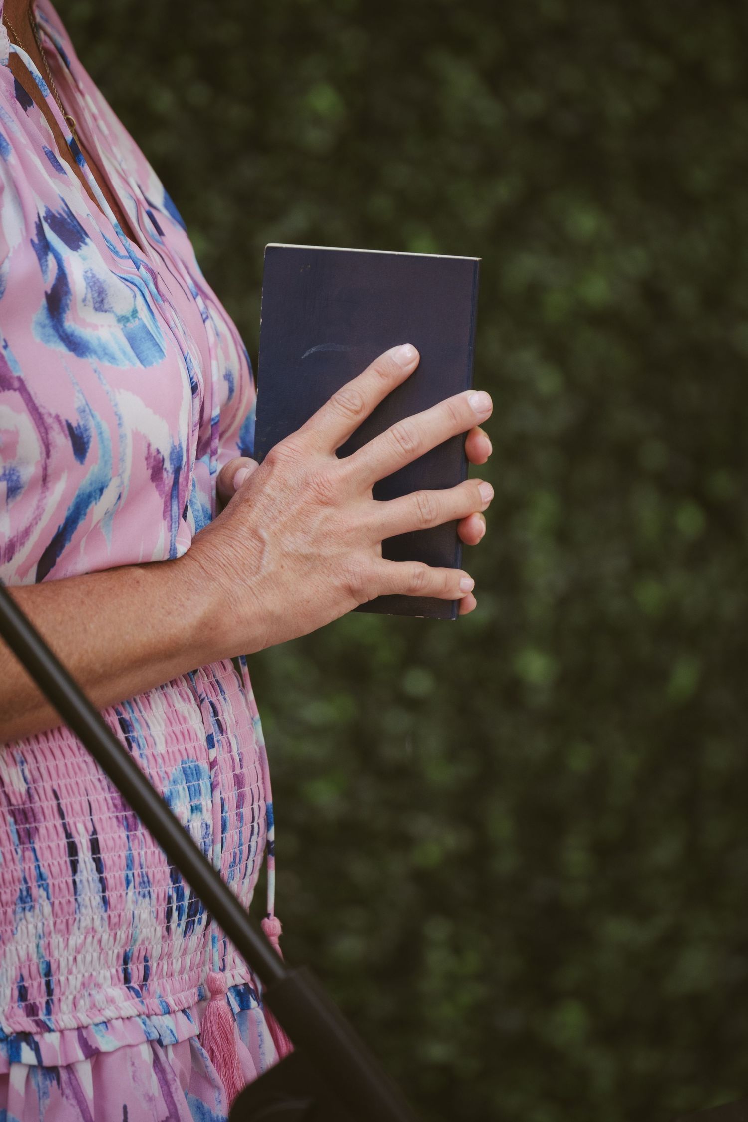 Woman in pink floral dress holds a navy book during an outdoor wedding ceremony.