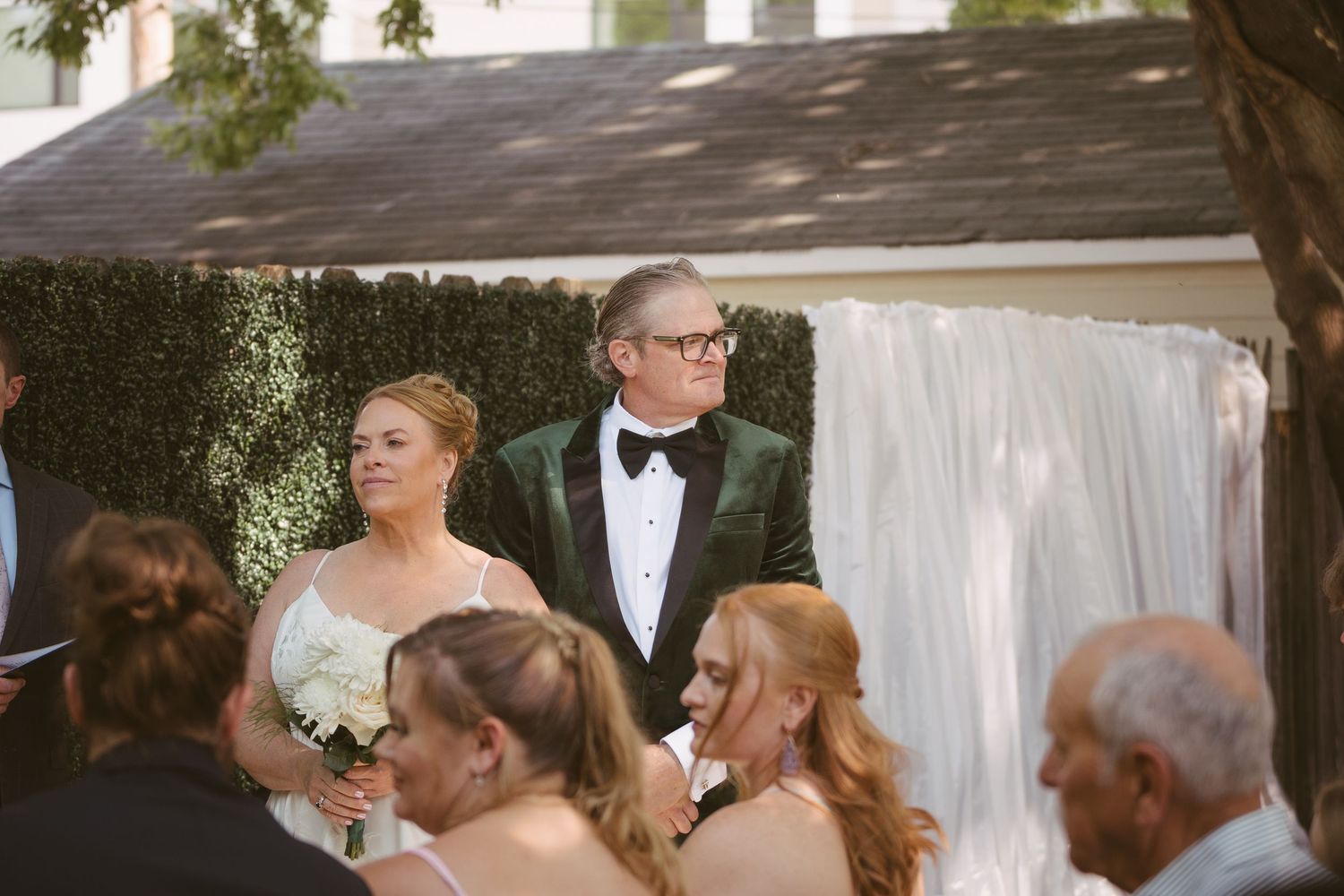 Bride and groom in green velvet tuxedo stand together during outdoor wedding ceremony with hedge backdrop.