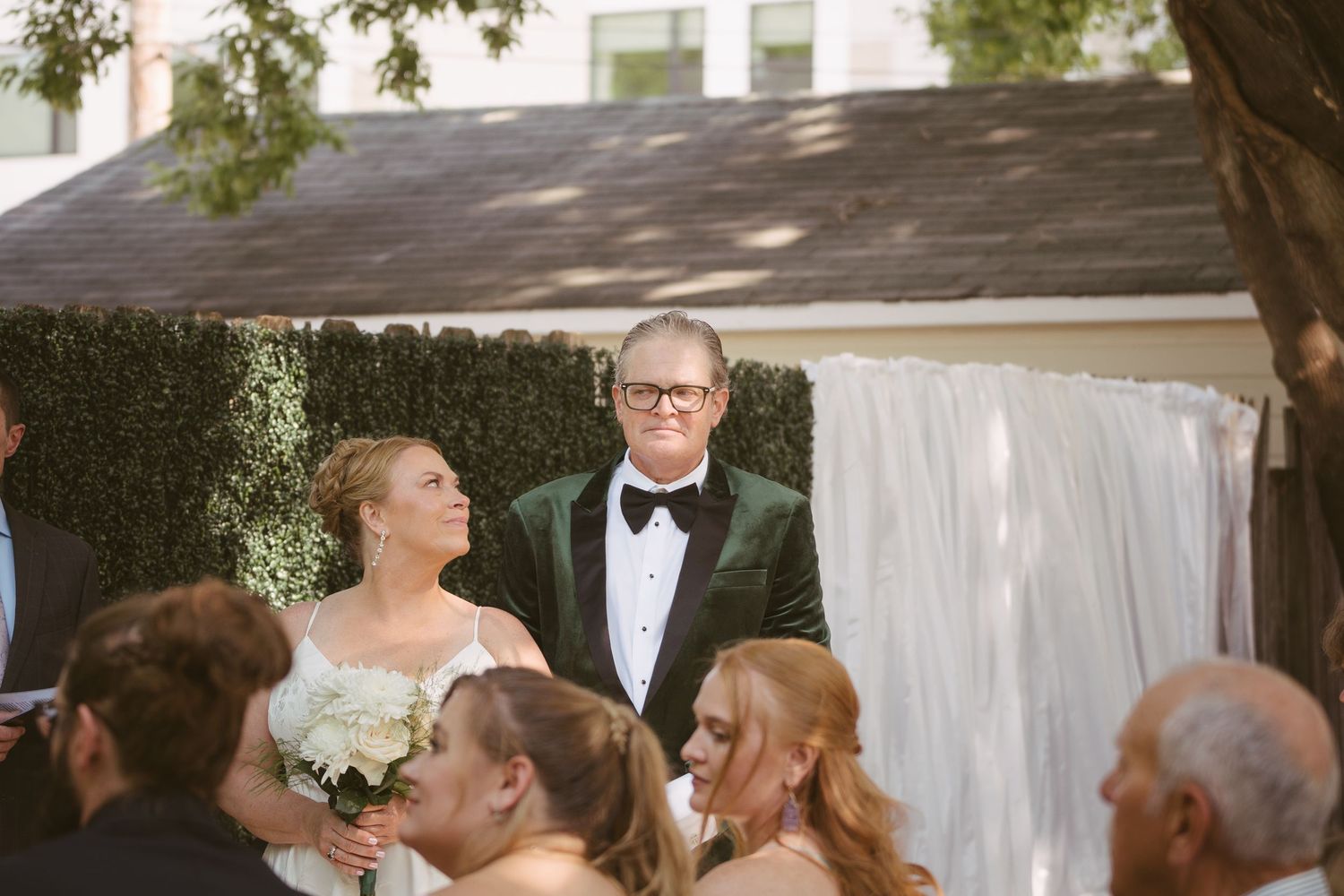 Bride holding white bouquet stands with groom in green velvet tuxedo at outdoor ceremony.