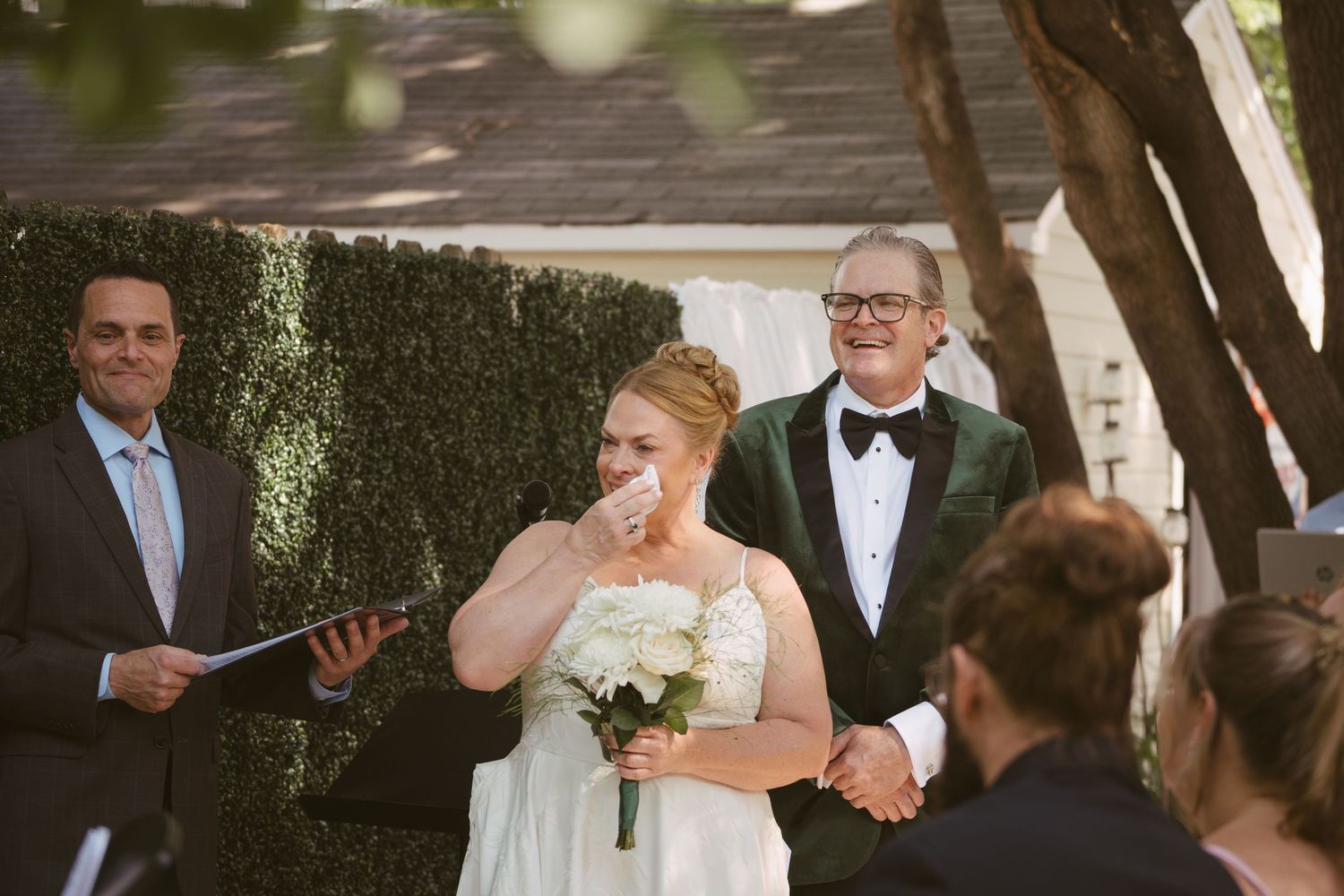 Emotional bride wipes tears while groom in green velvet tuxedo laughs at outdoor wedding.