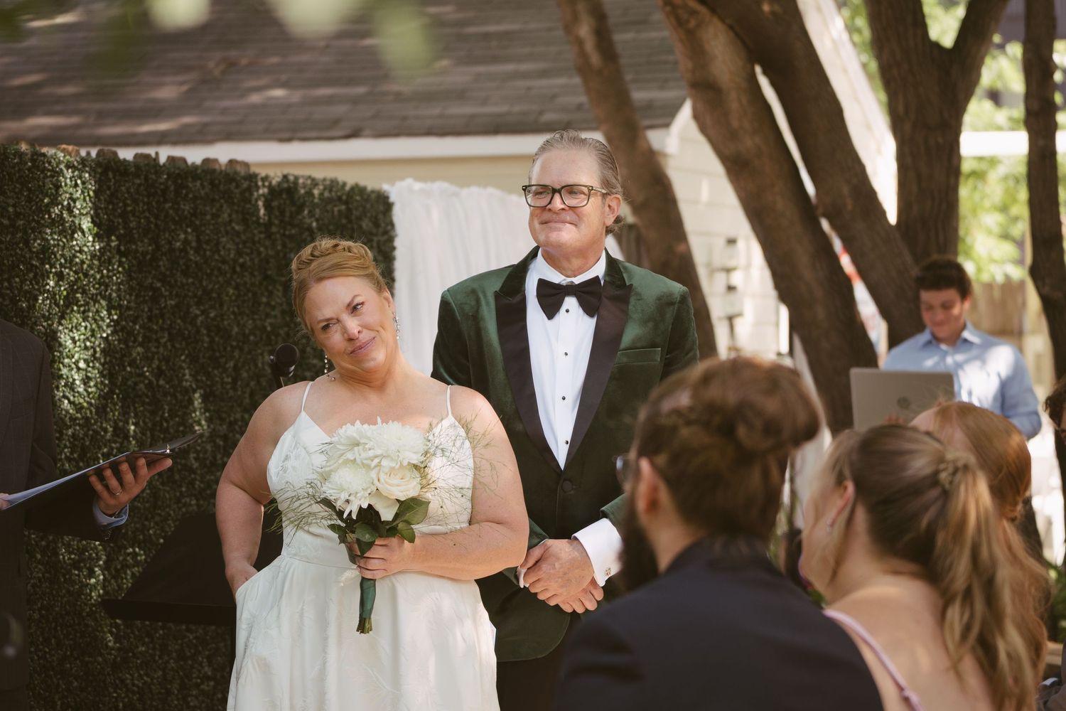 Bride holding white bouquet stands with tuxedo-clad officiant during outdoor wedding ceremony beside a green hedge wall.