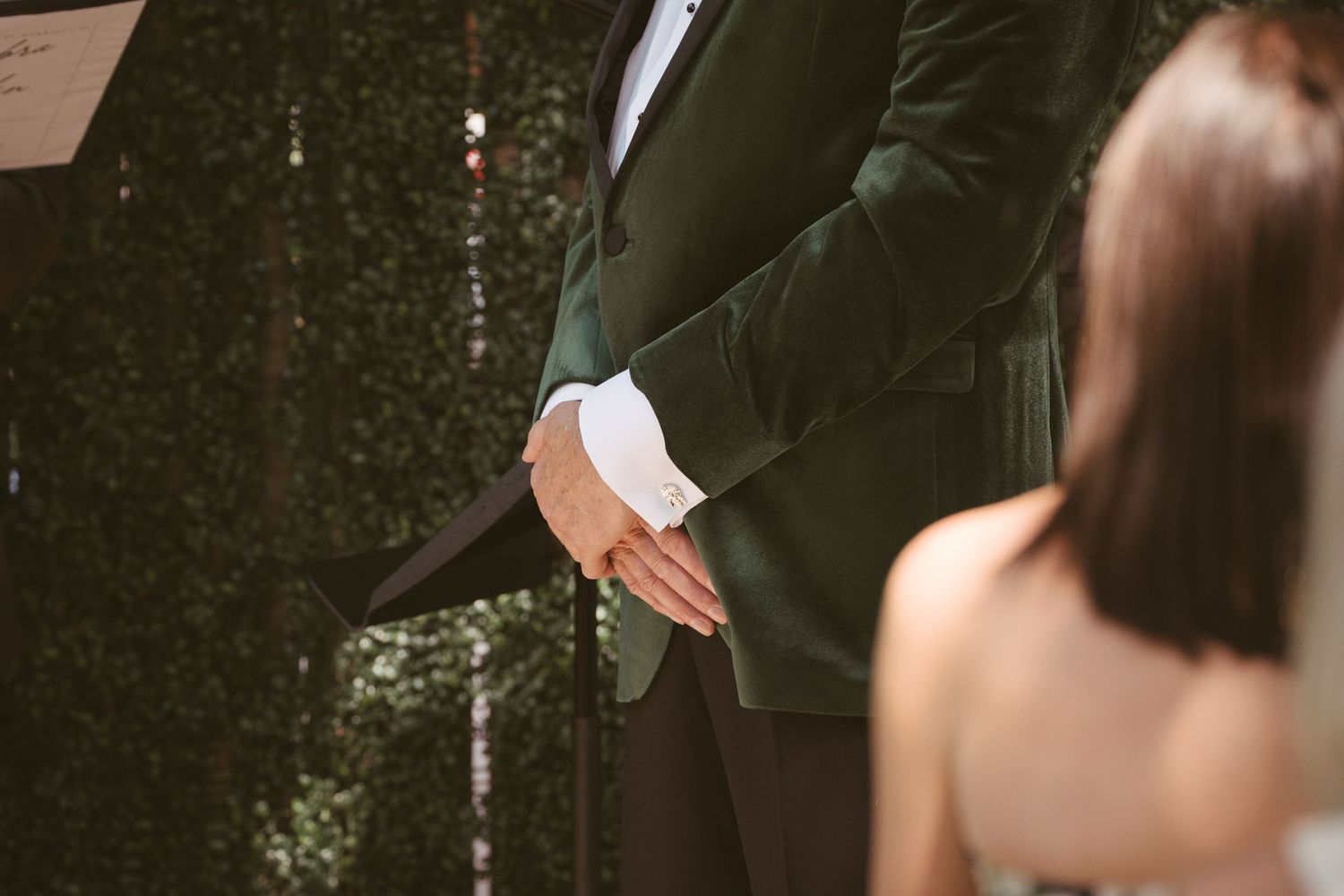 Groom in green velvet suit holding hands with bride in white dress at outdoor wedding ceremony.