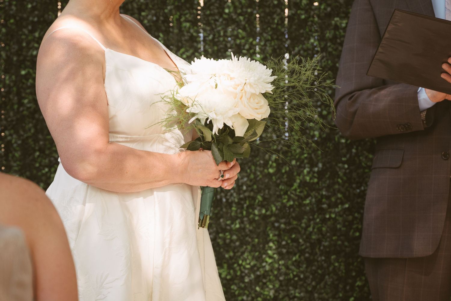 Bride in white gown holding white peony bouquet during outdoor wedding ceremony with green hedge backdrop.