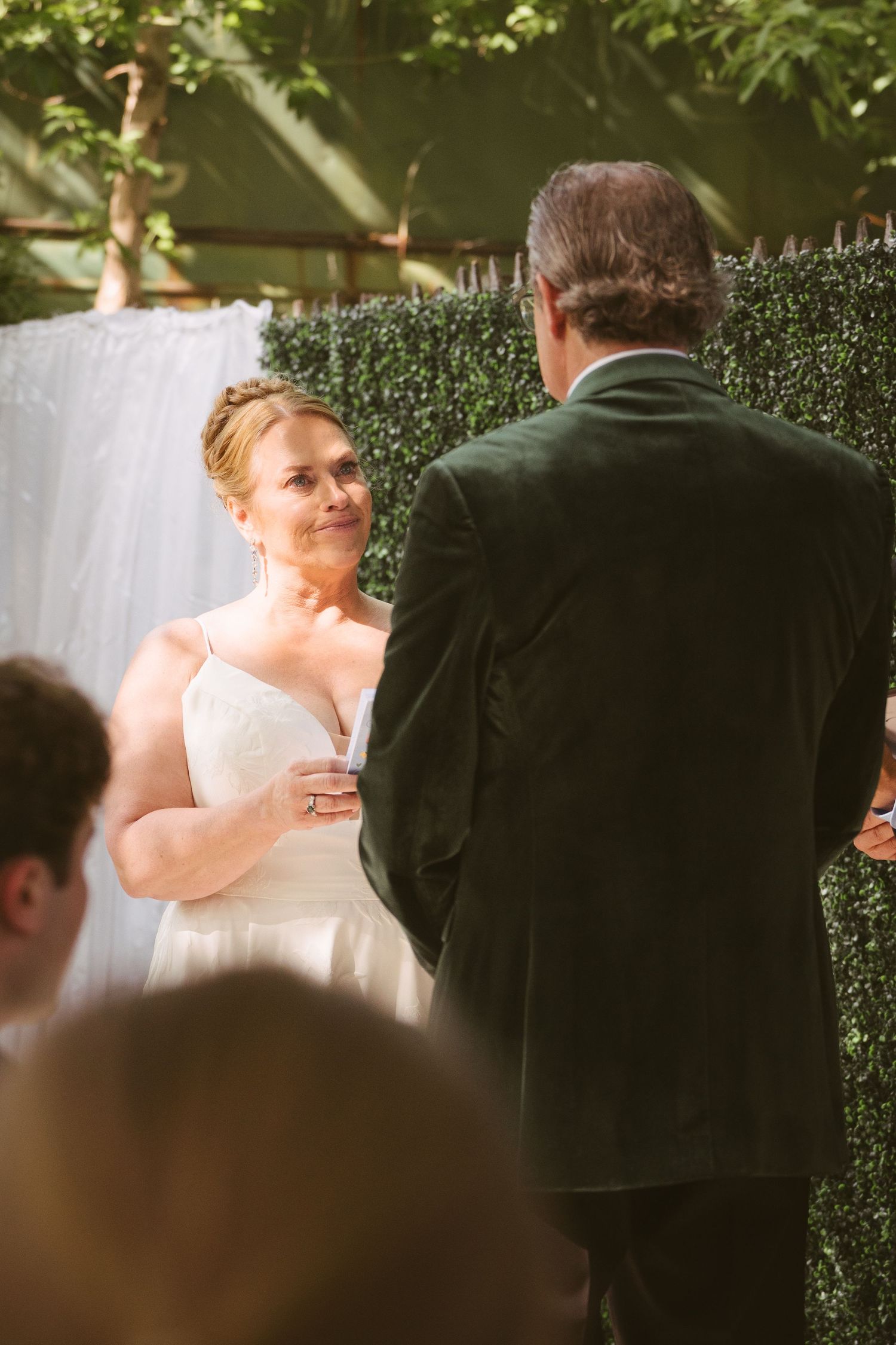 Smiling bride in white gown exchanges vows with groom in green velvet tuxedo at outdoor ceremony.
