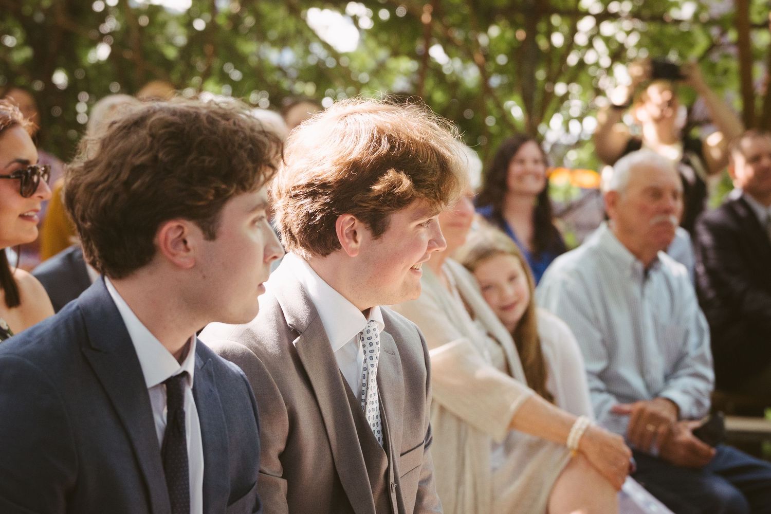 Young men in suits smile during an outdoor wedding ceremony surrounded by seated guests.