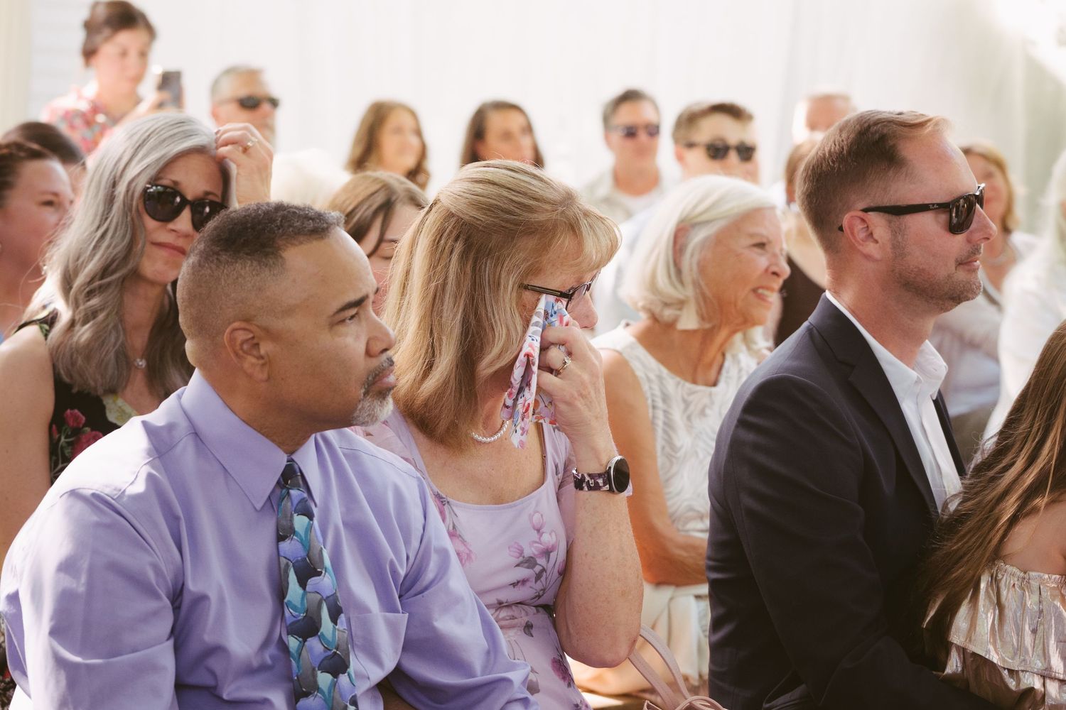 Wedding guests seated outdoors watching ceremony, woman wiping tears while man in purple shirt looks on.