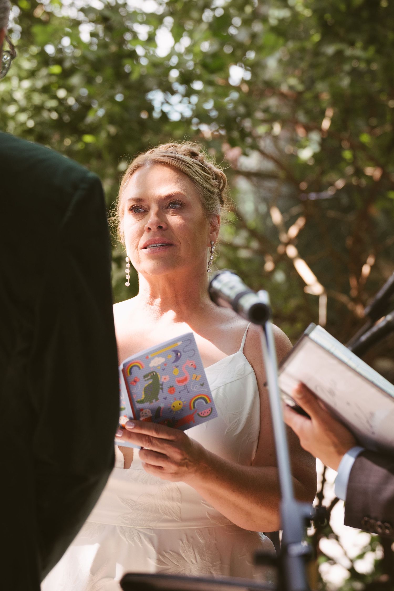 Bride holding colorful illustrated book during outdoor wedding ceremony, listening attentively at the altar.