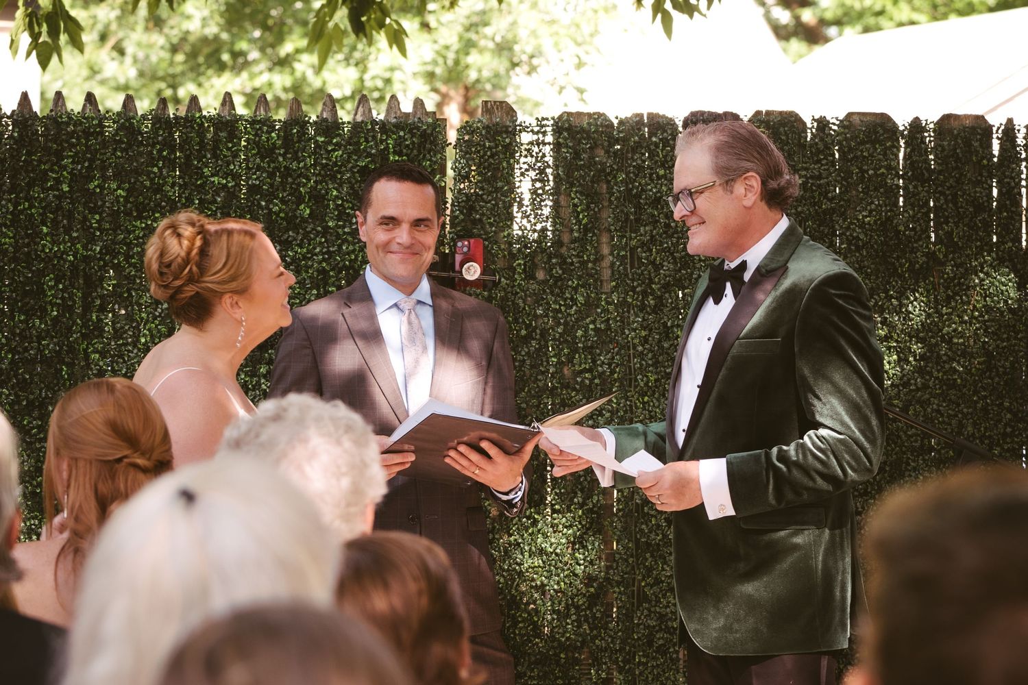 Wedding officiant in tuxedo reading vows to couple during outdoor ceremony by green hedge.