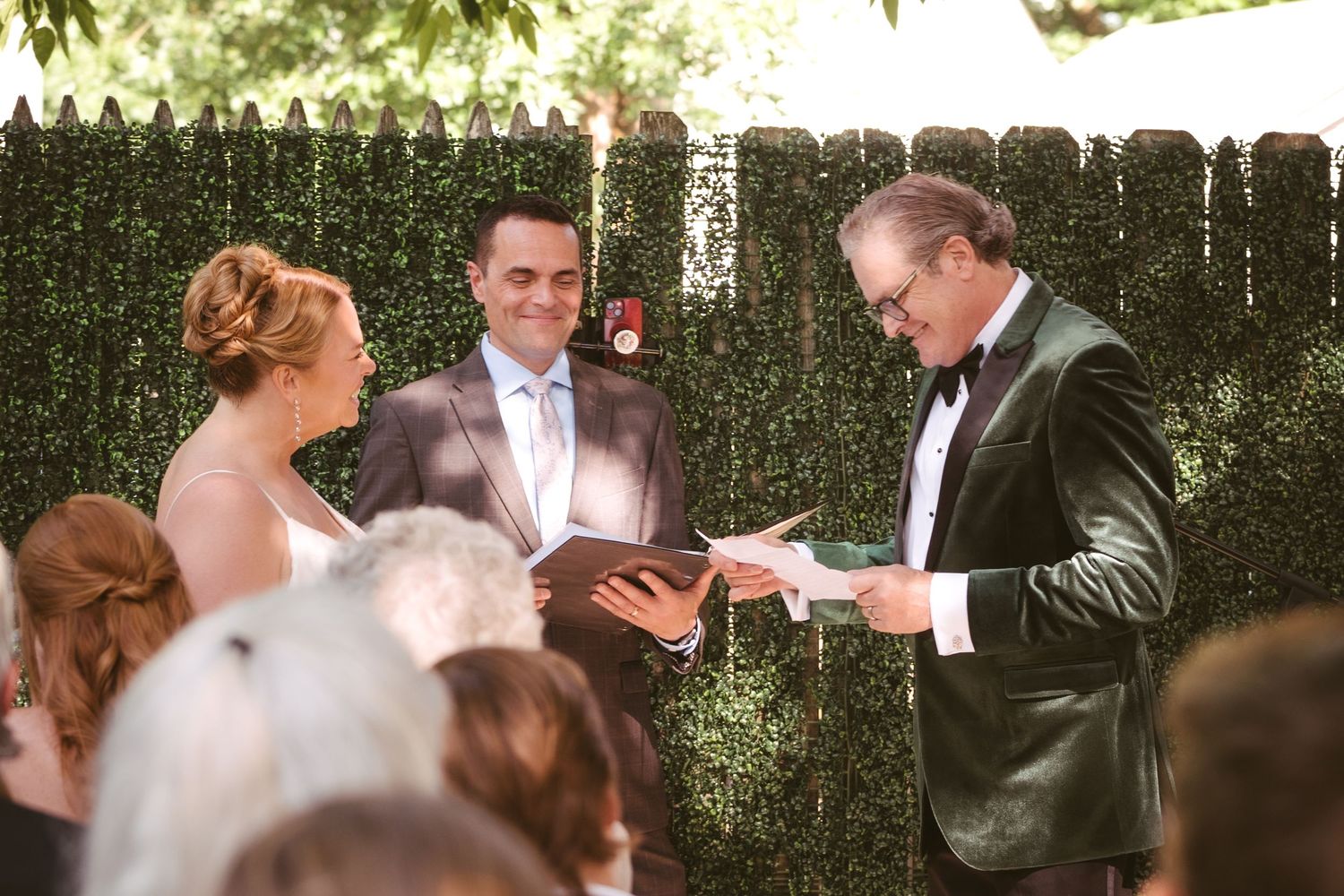 Bride and groom read vows with officiant during outdoor wedding ceremony against green hedge backdrop.