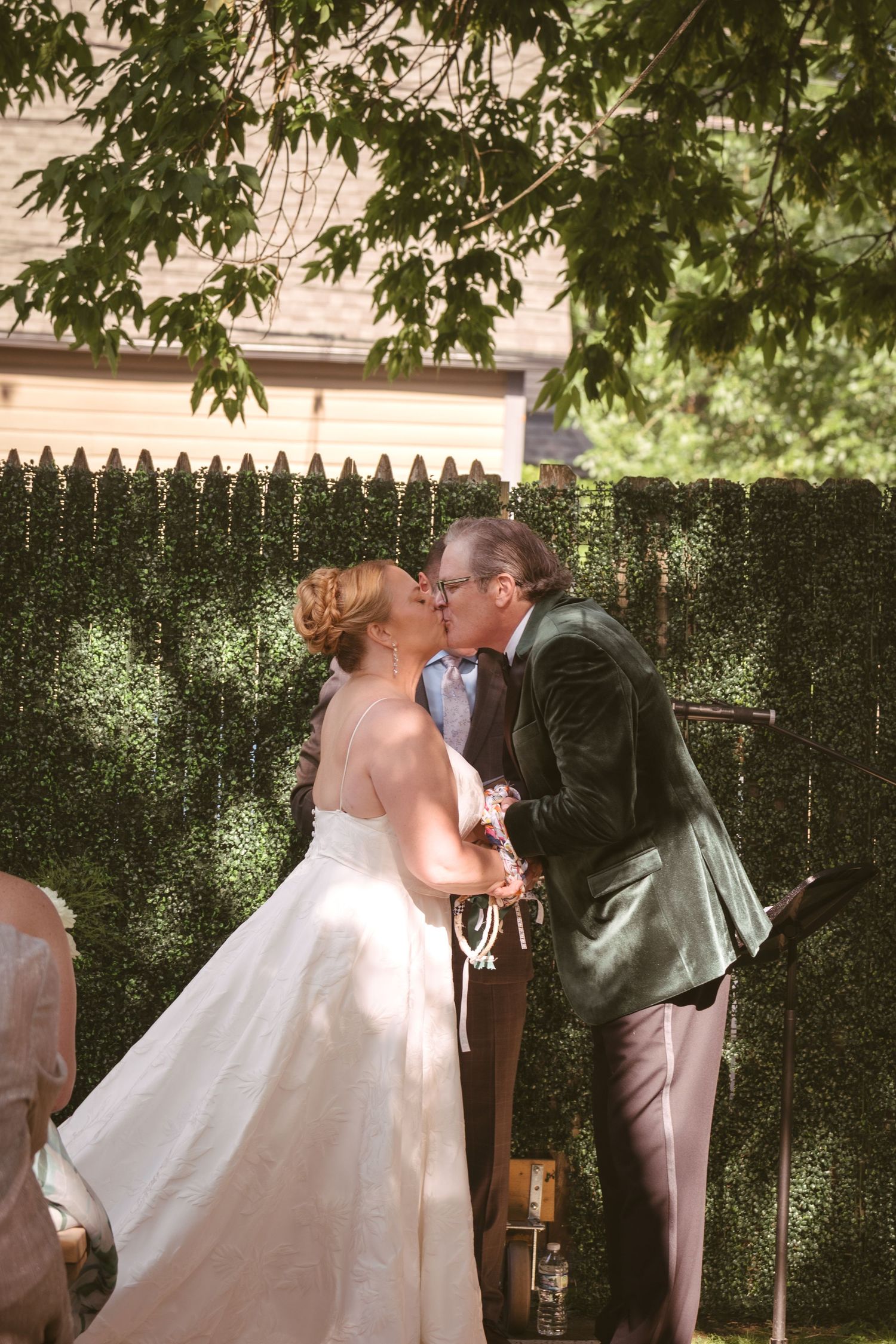 Bride and groom share their first kiss at an outdoor backyard ceremony before a lush green hedge wall.