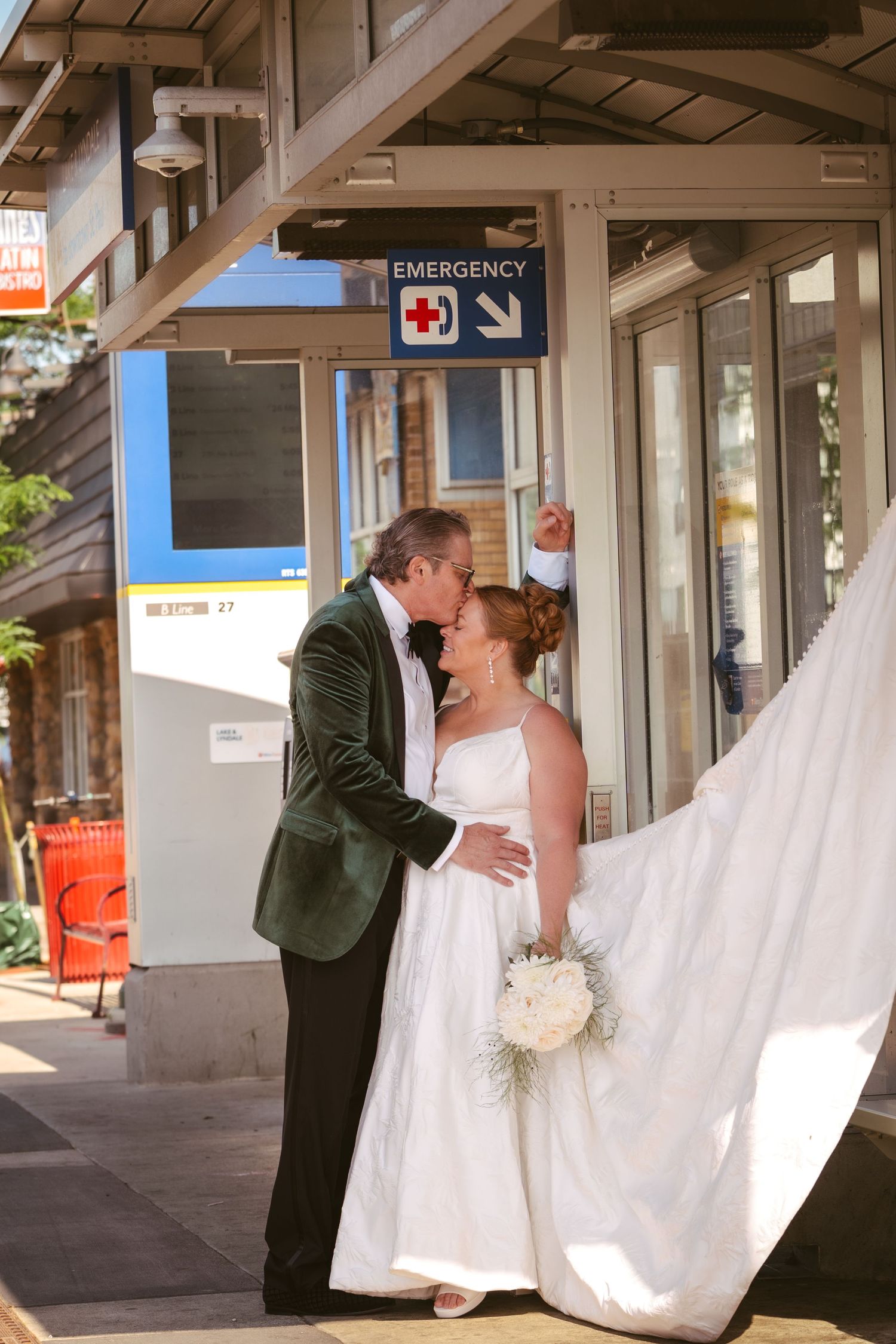 Bride and groom kiss outside near an emergency room entrance, bride's veil flowing in the breeze.