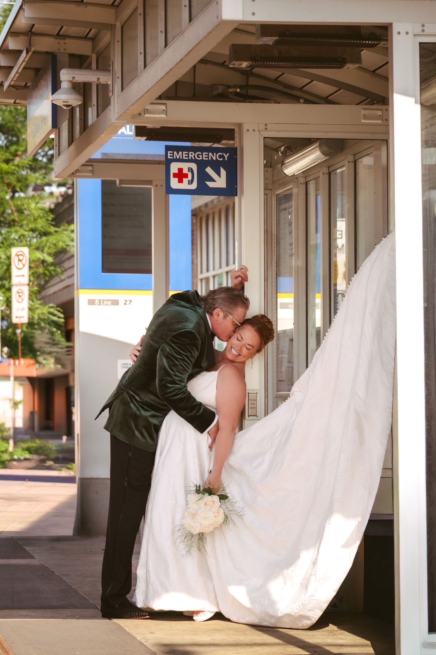 Newlywed couple sharing a romantic dip kiss outside a hospital emergency entrance, bride holding bouquet in wedding dress.