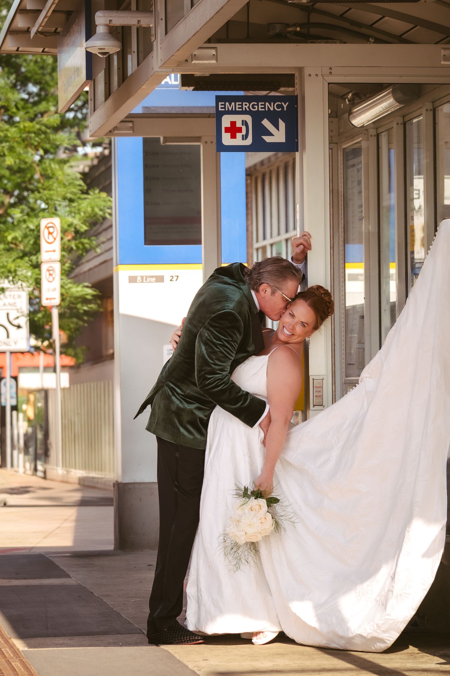 Groom in green velvet tuxedo dips bride in white gown outside a transit station emergency entrance.