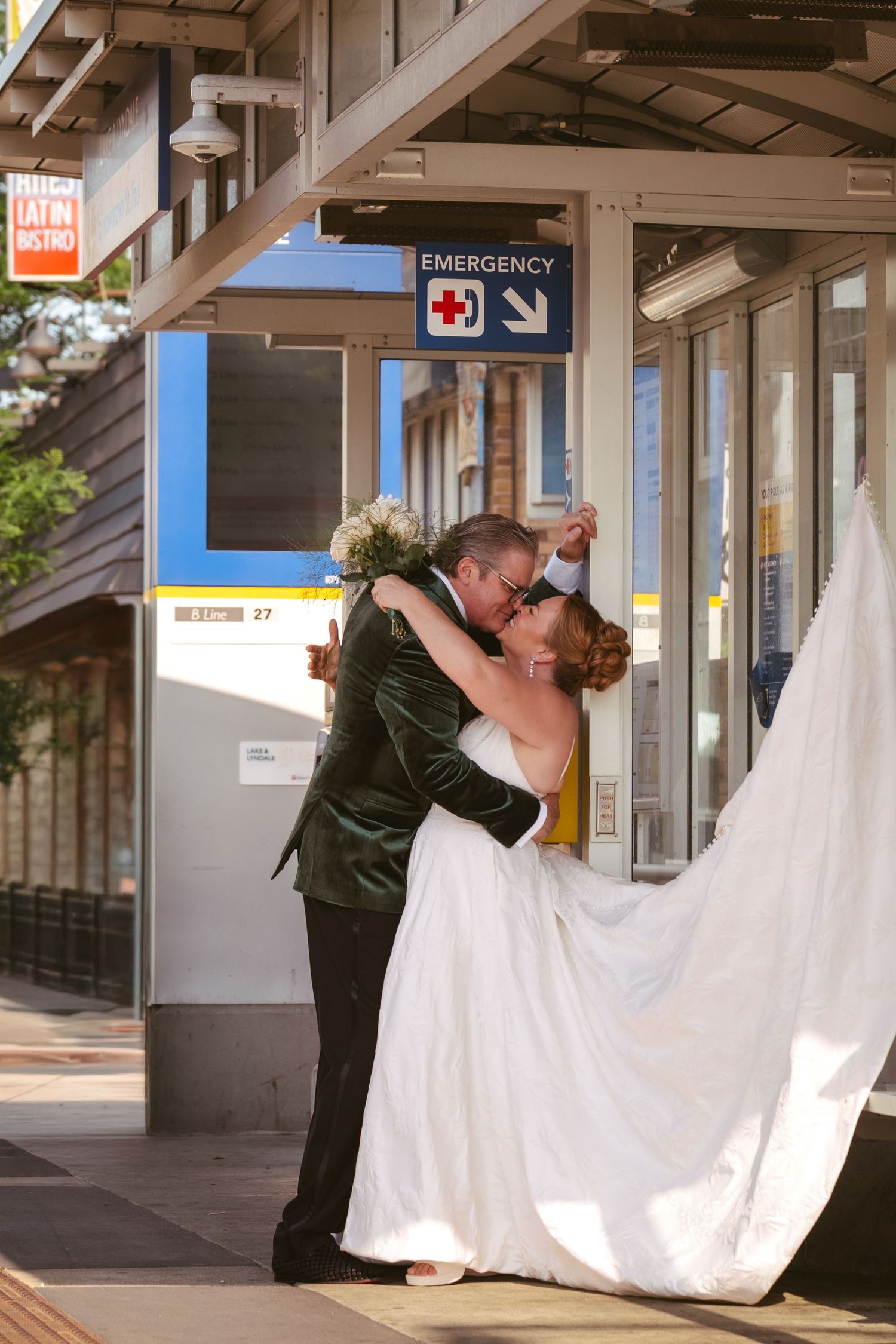 Groom dips bride in a romantic kiss at an outdoor transit station on their wedding day.