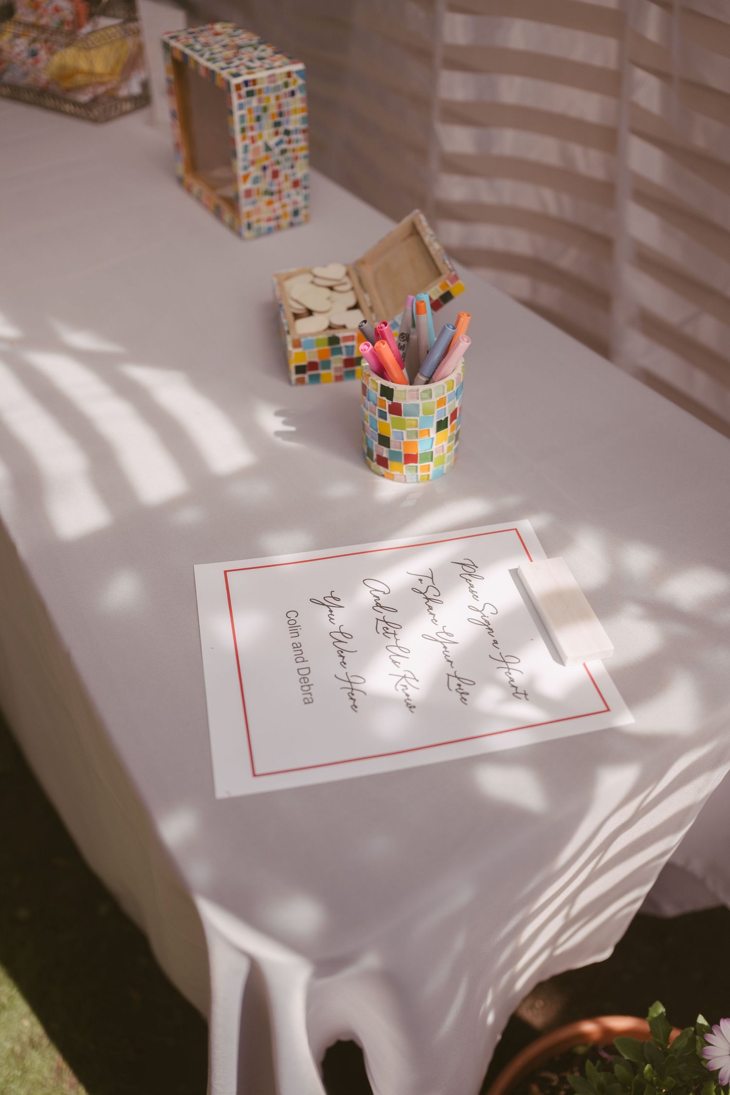 A white table with a colorful mosaic pencil holder, markers, and a calligraphy sign at an outdoor wedding.