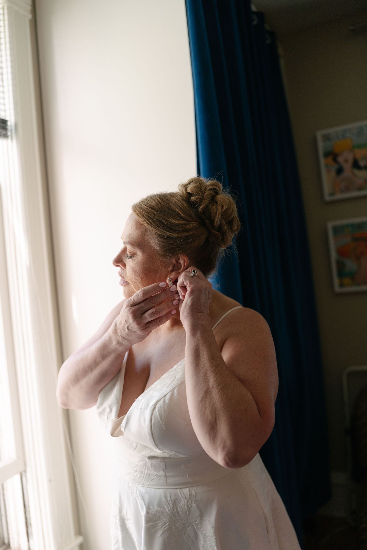A bride in a white wedding dress puts on earrings by a window with blue curtains.