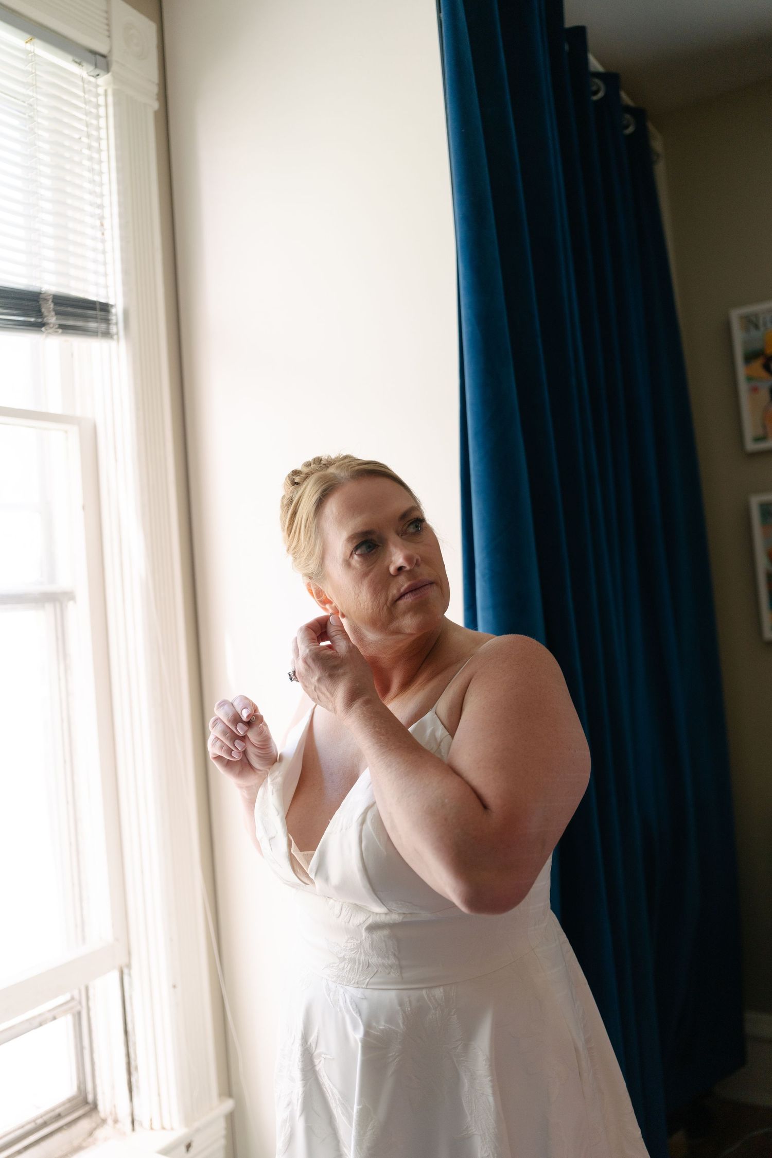 Bride in white wedding gown adjusting earring by window with blue curtains.