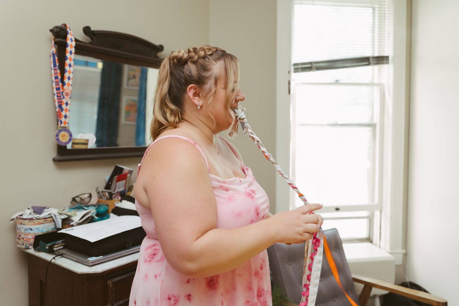 A woman in a pink floral dress holds a braided ribbon wand near a window indoors.