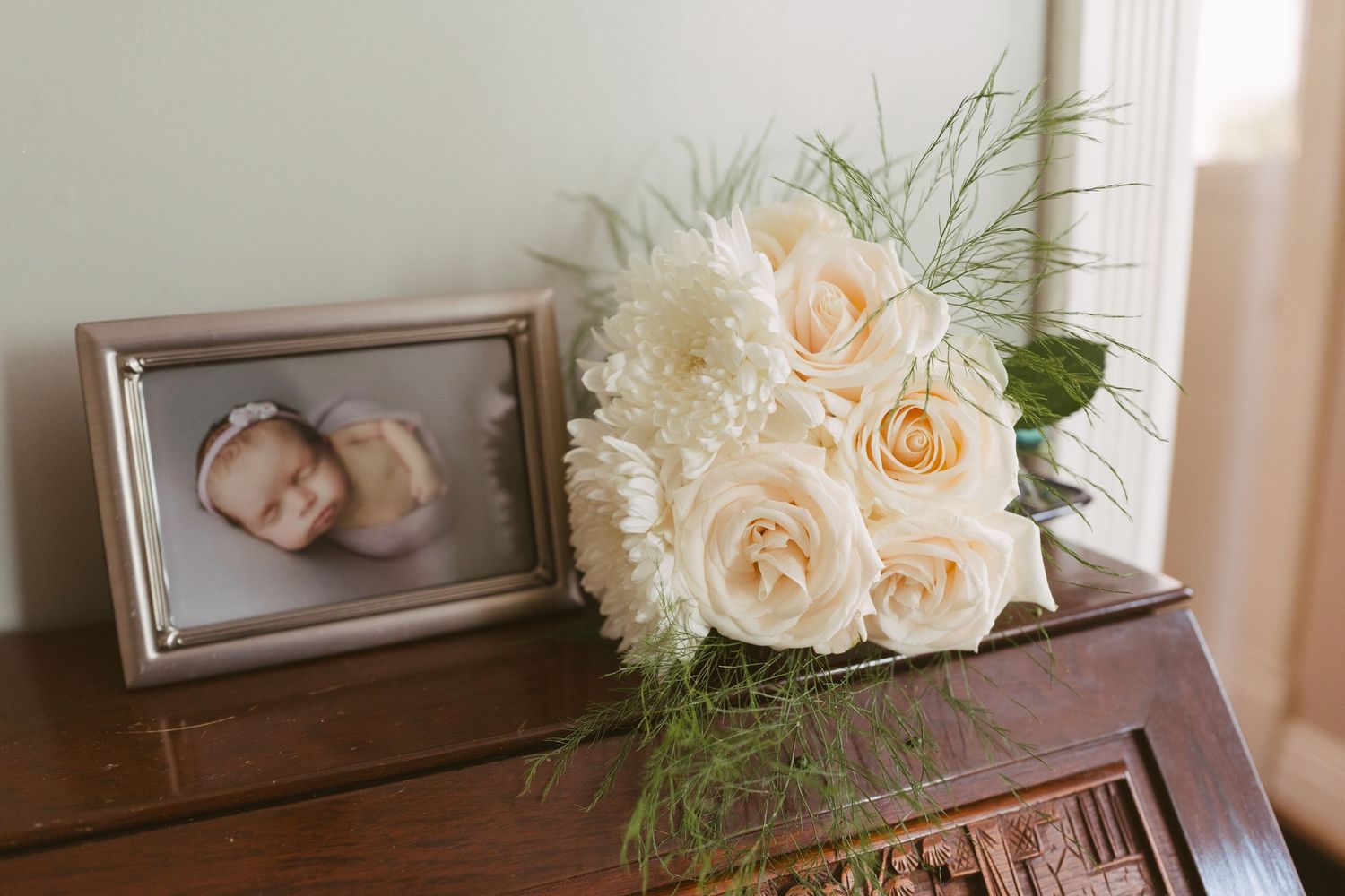 White rose and dahlia bridal bouquet beside a framed newborn photo on a wooden dresser.
